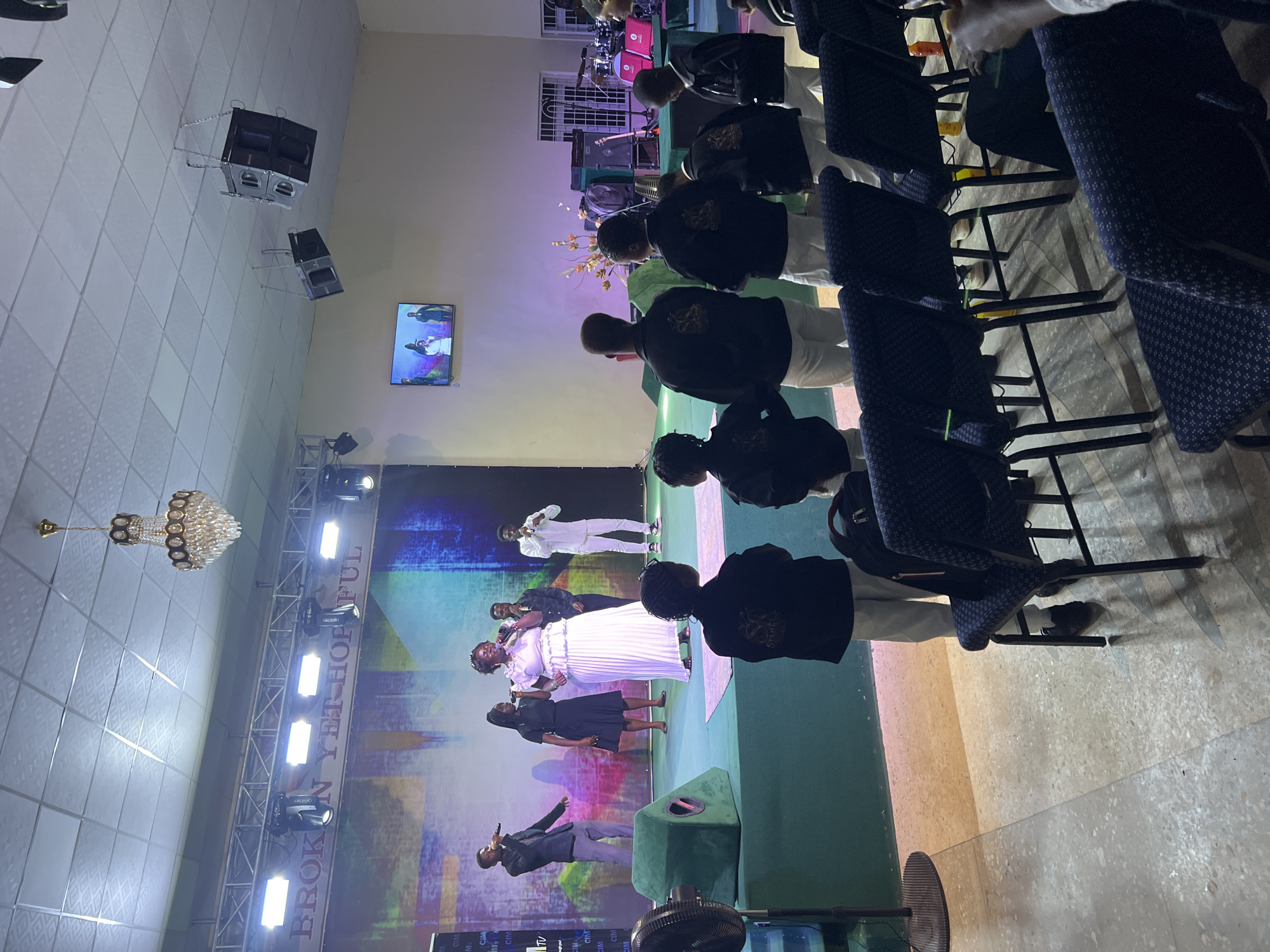 The image depicts an indoor event, likely a church service or performance, taking place in Gura Top, Nigeria. A group of performers, including singers and a woman in a white dress, are on a raised stage with a colorful backdrop that reads "BROKEN YET HOPEFUL". Several children, dressed in matching black jackets and white pants, are standing in the foreground, facing the stage. They appear to be part of a choir or performance group. The environment is illuminated by stage lighting and a chandelier hanging from the ceiling. There are also speakers mounted on the ceiling and on the stage. In the background, musical instruments and a drum set are visible. The audience is seated in dark blue chairs, some of which are visible in the foreground and to the right of the image. The floor is a terrazzo-like material.