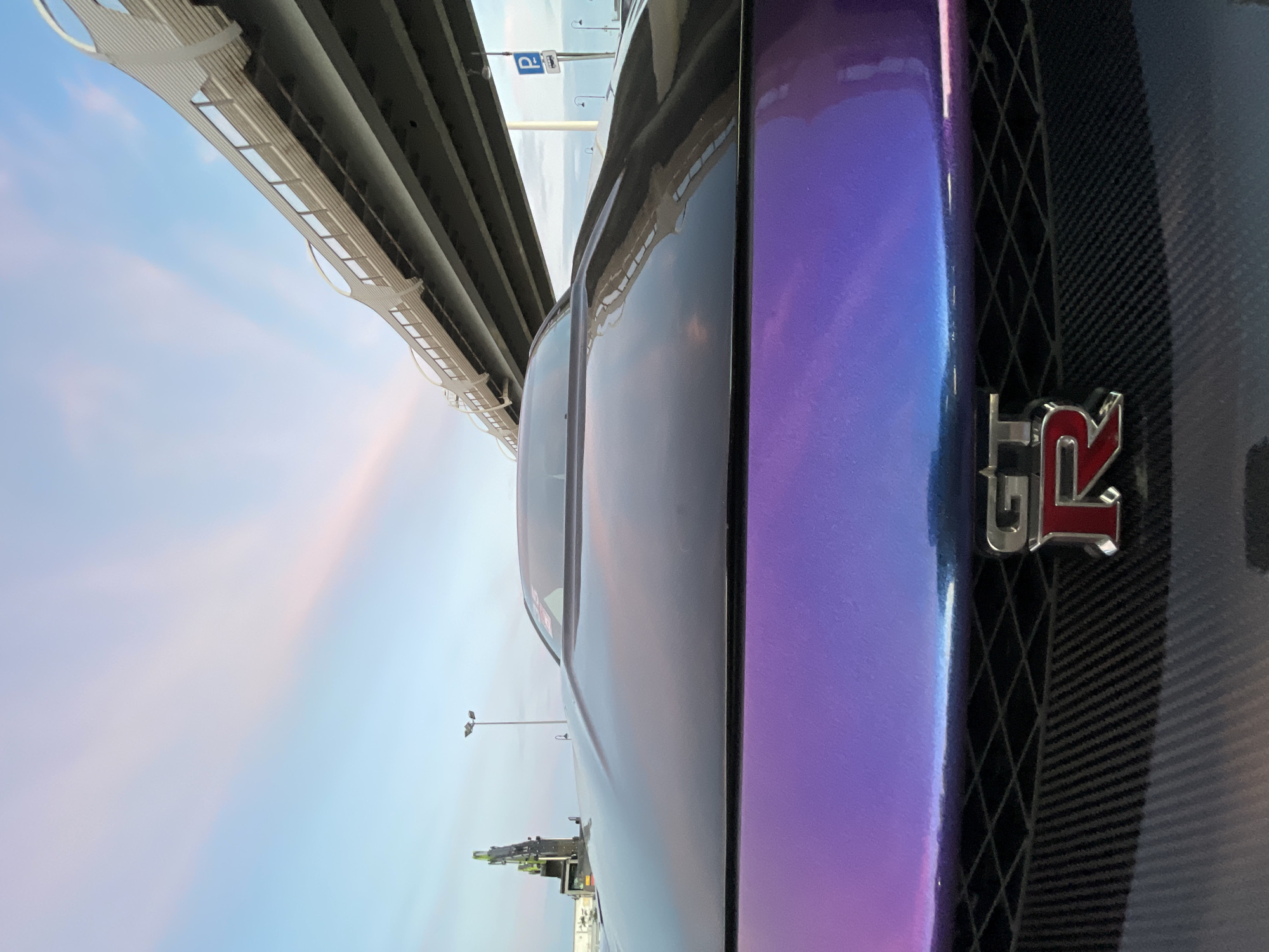 A low-angle, close-up shot focuses on the front of a black and purple car, likely a Nissan GT-R given the prominent "GT-R" badge. The car's hood has a sculpted center bulge and reflects the sky, which is a gradient of pale blue to pink from the sunrise or sunset. In the background, a large overpass or bridge structure dominates the upper right of the frame, casting shadows. To the left, a construction crane with a yellow boom is visible. A streetlamp and a parking sign are also present near the bridge. The overall environment appears to be an urban or semi-urban area, possibly near a highway or industrial zone.