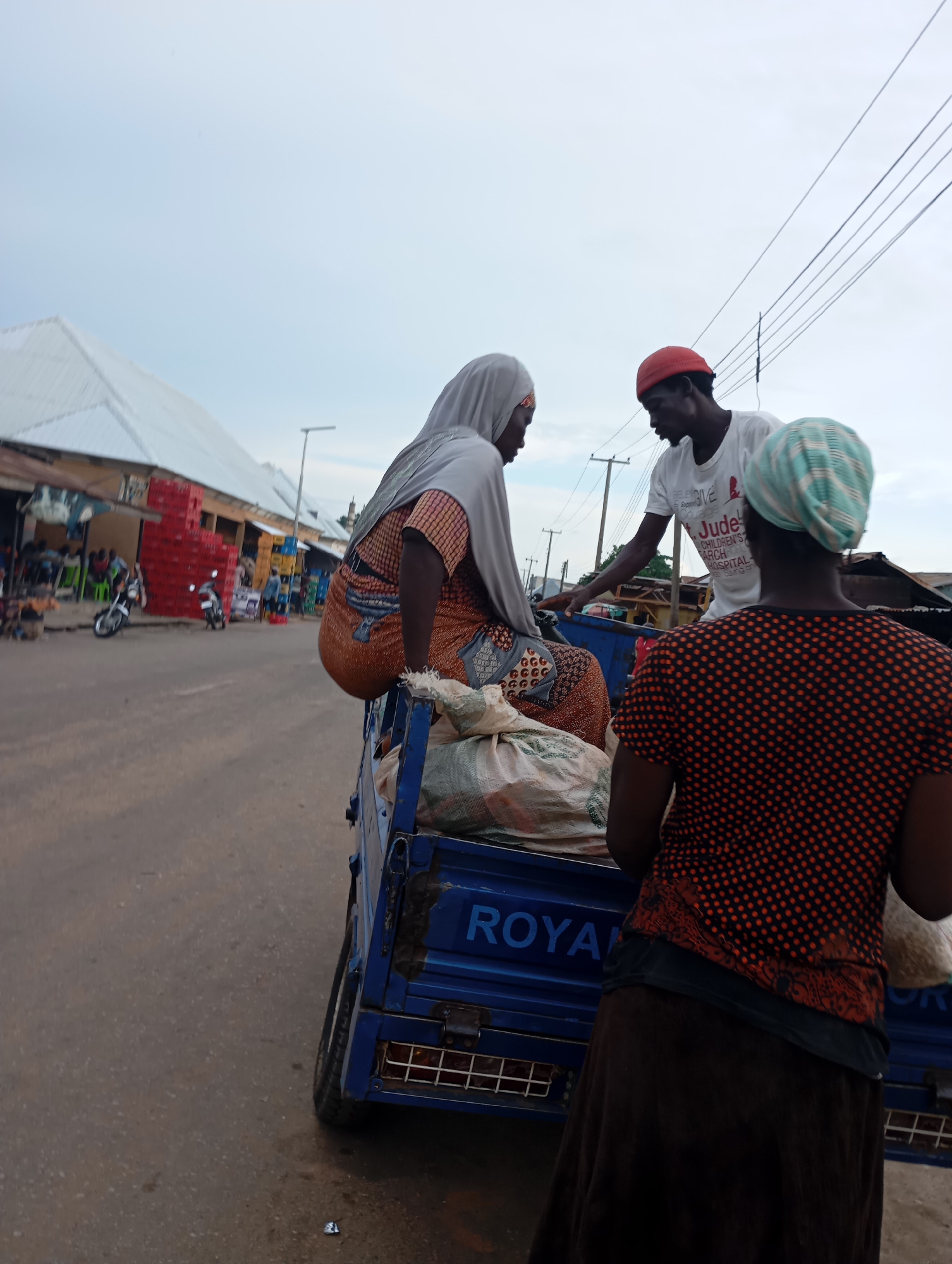 In Shendam, Nigeria, a low-angle, medium shot captures three individuals near a blue truck. A person wearing a red cap and white t-shirt, with text that appears to read "BELIEVE GIVE Courage Jude Children's Hospital," stands in the truck, engaging with two women. One woman, seated in the truck, wears a light grey headscarf and an orange and white patterned garment. The other woman, standing and facing away, has a light green and blue striped headscarf and wears an orange and black polka dot shirt over a dark skirt. The truck bed contains several large sacks of produce. The background depicts a street scene with market stalls featuring red and yellow crates, a motorcycle, and a few people in the distance. The sky is overcast.
