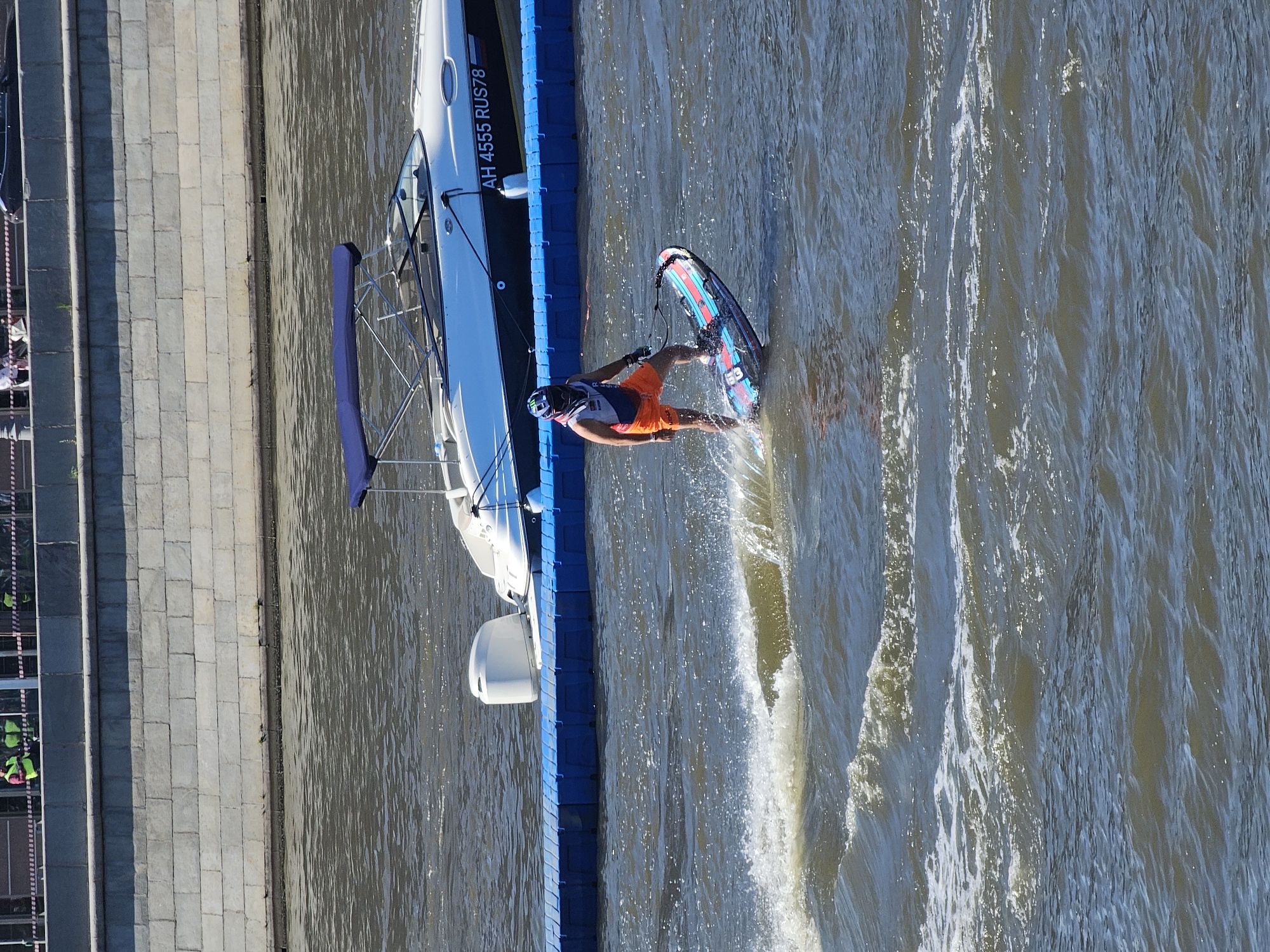 An overhead shot shows a person riding a jet ski on a body of water, with a large boat docked on a blue pontoon in the background. The person on the jet ski is wearing orange shorts, a white and blue life vest, and a helmet. They are actively riding, creating a wake behind them. The boat is white with a blue canopy and has the registration number "AH 4555 RUS78" printed on its side. A stone wall lines the edge of the water, with a walkway or promenade above it. The overall scene appears to be an outdoor recreational area within a city.