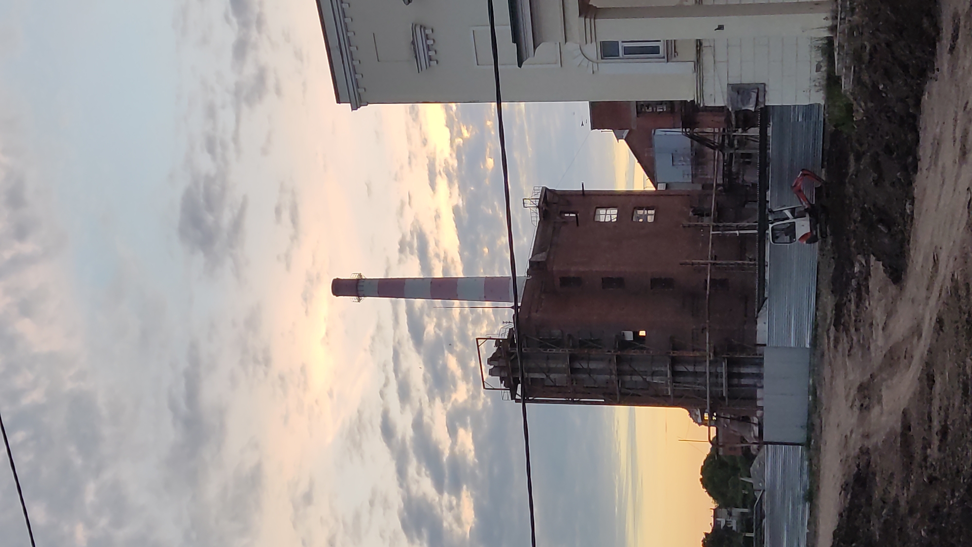 A brick industrial building with a red and white striped smokestack stands in the center of the frame, against a cloudy sky. The building appears to be under construction or renovation, with scaffolding visible on its side. To the right, a portion of a cream-colored building with classical architectural elements is visible. In the foreground, there is a construction site with dirt piles and a small excavator. The image appears to be taken in Krasnodar, Russia, given the architectural style and the presence of industrial elements.