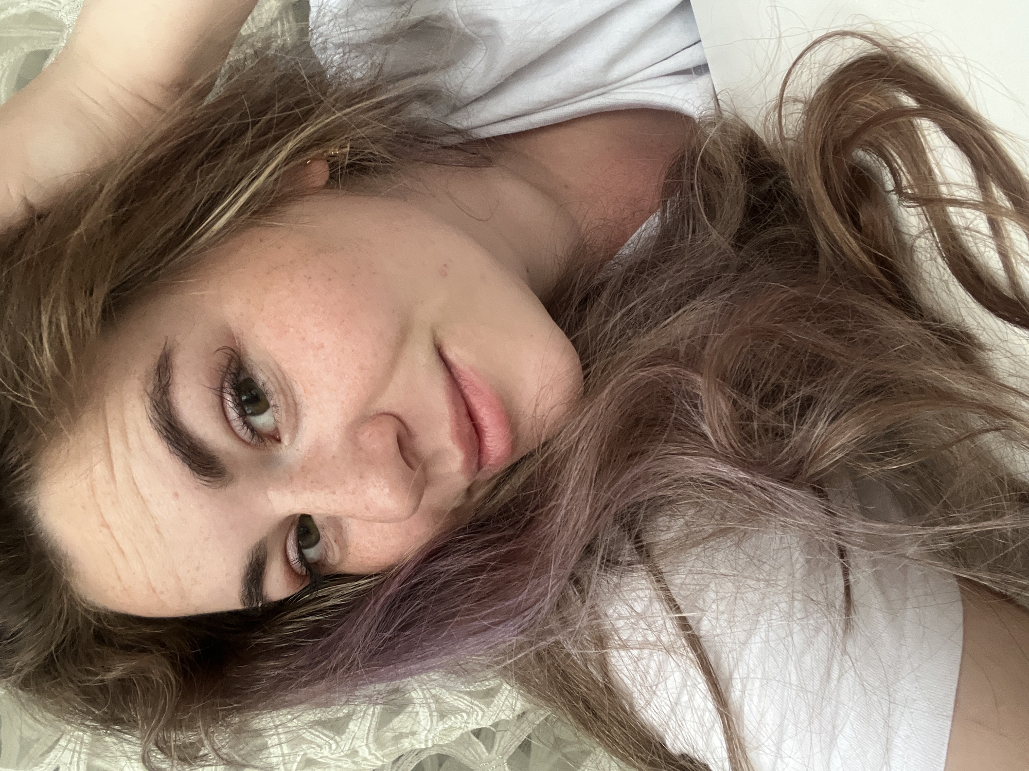 A selfie-style portrait depicts a Caucasian woman with light skin, freckles, and light brown hair with hints of purple. She is lying down, her head angled towards the viewer. The visible clothing is a white t-shirt. The background appears to be a white surface with a textured pattern, possibly a bedspread or similar fabric. The specific location in the city of Chaikovsky, Russia cannot be determined from the image.