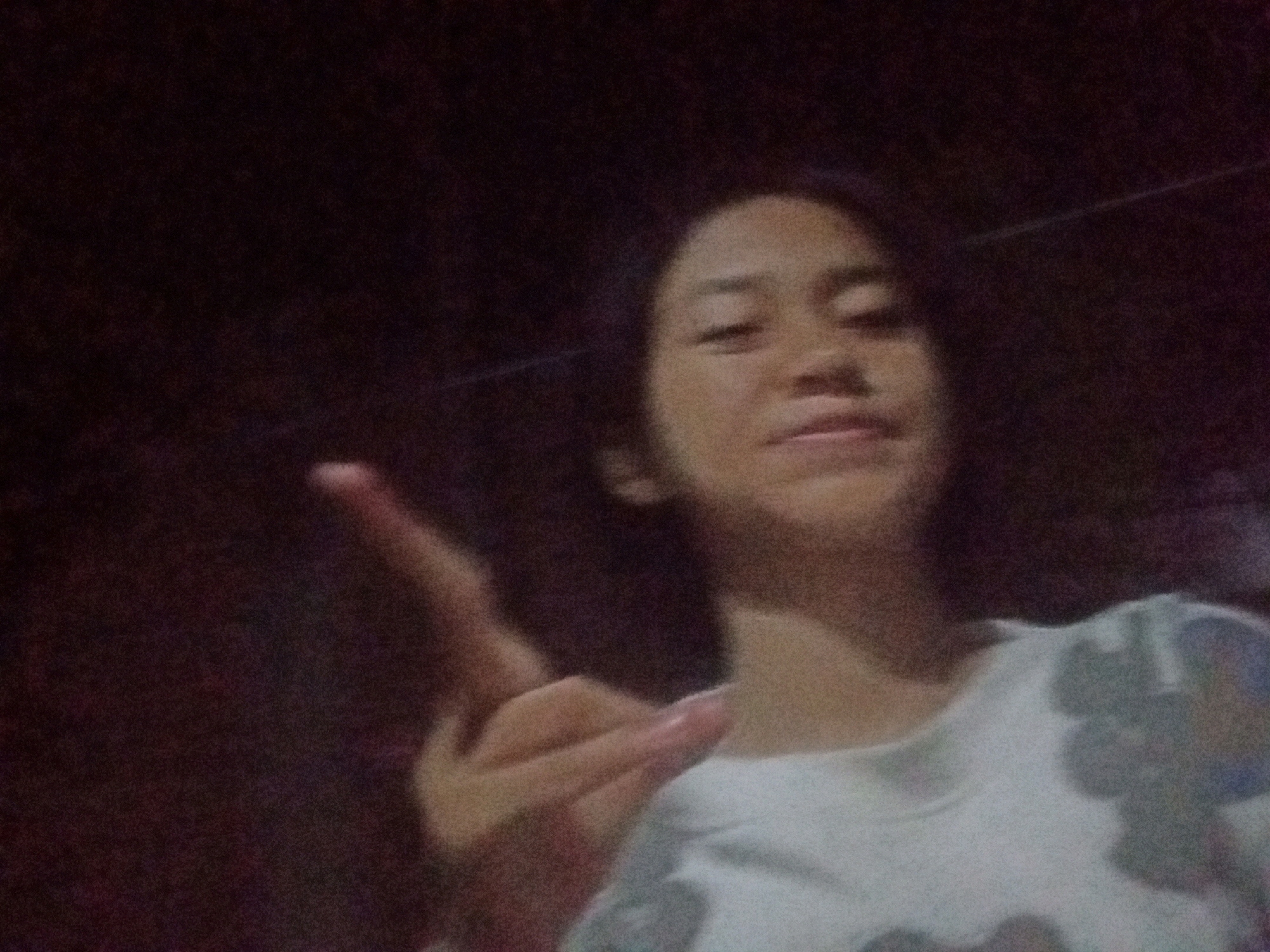 A young woman is taking a selfie in what appears to be a dimly lit room. She is wearing a white shirt with a floral pattern. She is making a peace sign with her left hand, which is positioned in the lower left quadrant of the image. Her face is partially visible in the upper right quadrant. The background is dark and out of focus, suggesting a low-light environment. The image appears to be taken indoors, possibly in a residential setting. There is no specific indication of the city of Badas, Indonesia, in the image itself.