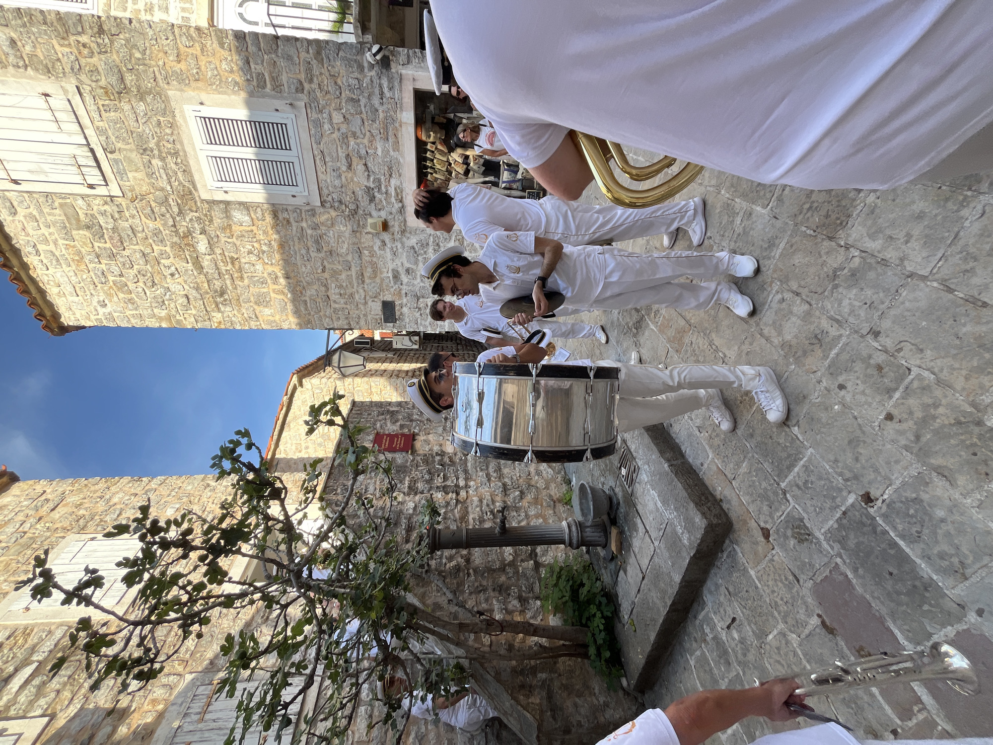 A street-level, medium shot depicts a musical band in white uniforms parading along a narrow, cobbled street, likely in the old town of Budva, Montenegro. The band members, wearing sailor-style hats and playing various instruments including a large drum, a tuba, and a trumpet, are seen from a slightly low angle.
The background shows stone buildings with windows and architectural details. A tree with leaves and branches is visible, adding a touch of greenery to the urban setting. The sky is visible above the buildings. The overall scene appears to be a cultural event or performance taking place in the historic part of the city.