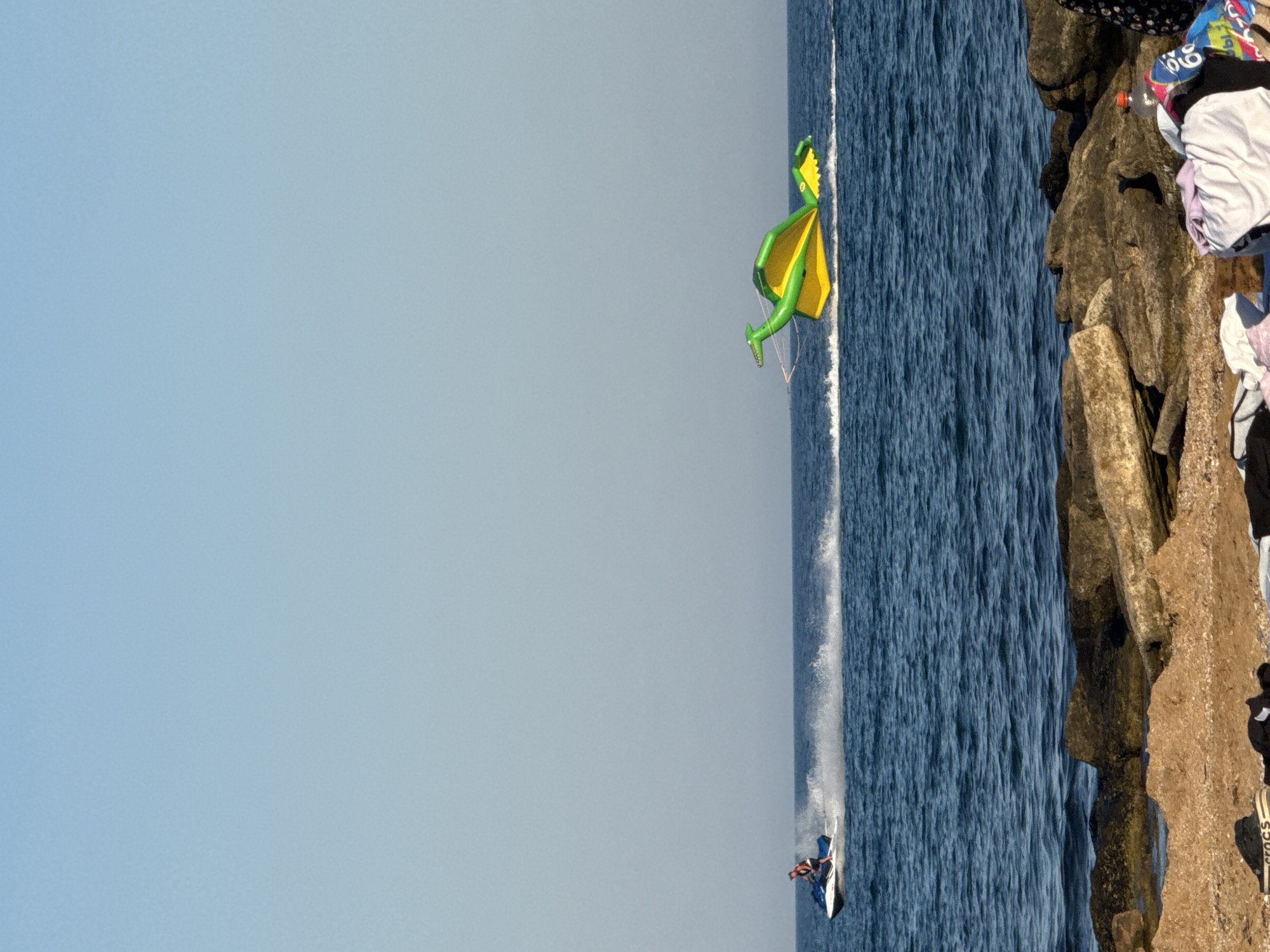 In an outdoor scene with a clear blue sky and dark blue water, a person rides a white jet ski creating a spray of water as they move across the sea. To the right, a large, inflatable yellow and green dragon-shaped float is being towed behind an unseen vessel, also creating a wake. The foreground features a rocky beach with scattered large stones and sand, with some clothing and a plastic bottle visible on the ground. The scene appears to be set in a coastal area, likely a beach resort, although the specific location in the city of Unknown, Ukraine cannot be determined from the image alone.