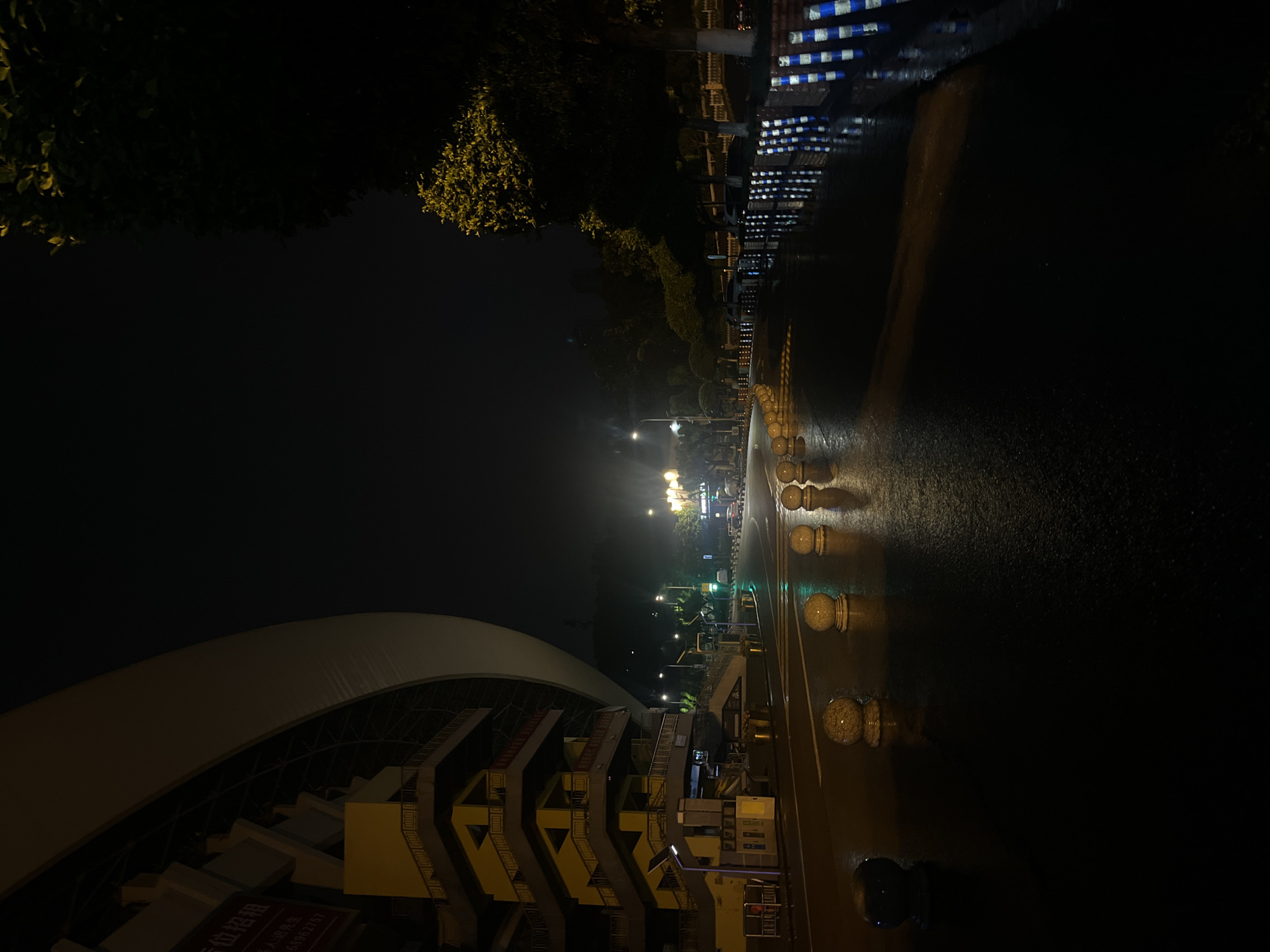 This image captures a nighttime urban streetscape in the city of Jiulongpo District, China, characterized by wet asphalt reflecting artificial light sources. On the left, a large, modern building with a prominent curved, light-colored upper facade dominates the frame. Below this, a multi-story section features yellow-ochre exterior walls and dark railings, forming a series of geometric outdoor staircases. A partially visible sign on the building displays Chinese characters, including "位招租" (spaces for rent) and a phone number "639962757."
The foreground and midground are dominated by a dark, wet road surface, indicating recent rainfall. A line of light-colored, spherical bollards with reflective bases runs along the edge of the road, separating it from the adjacent areas. Further down the right side of the road, blue and white striped roadside barriers are visible, also reflecting light. Trees with green foliage, some illuminated by streetlights with a yellowish glow, line the right side of the street. In the distance, multiple streetlights cast pools of light, and a traffic light displays a green signal. Faint outlines of vehicles can be discerned further down the road. No individuals are visible in the immediate scene.