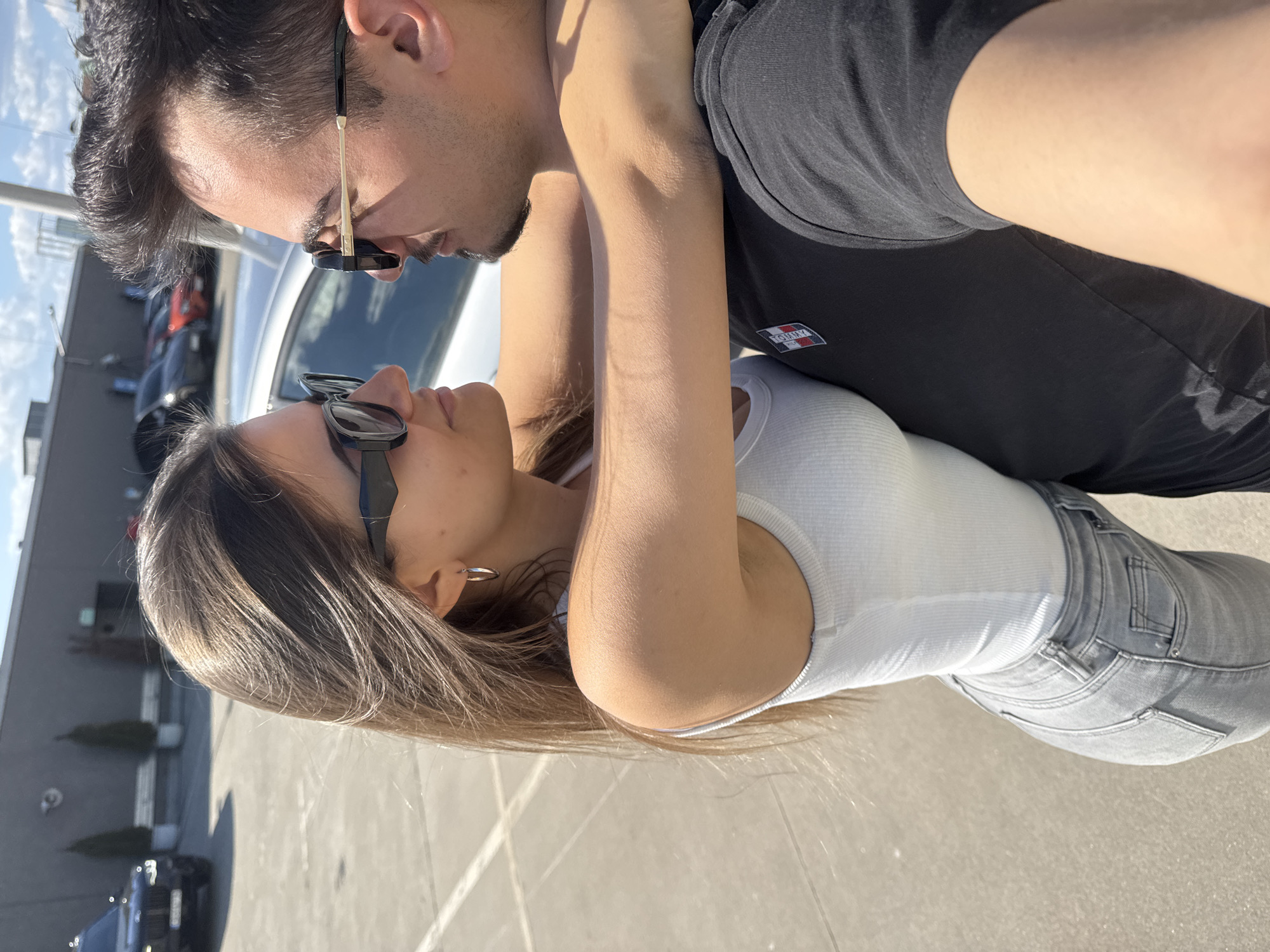 A man and a woman are embraced in a parking lot. The man is wearing sunglasses and a black t-shirt with a "Tommy" patch logo on the upper left. The woman wears sunglasses, a white ribbed tank top, and grey jeans. They are looking at each other. Vehicles are in the background, including a white car and black SUV. The location is in a city that may be Sofia, Bulgaria, based on clues found in previous turns. The time is daytime.