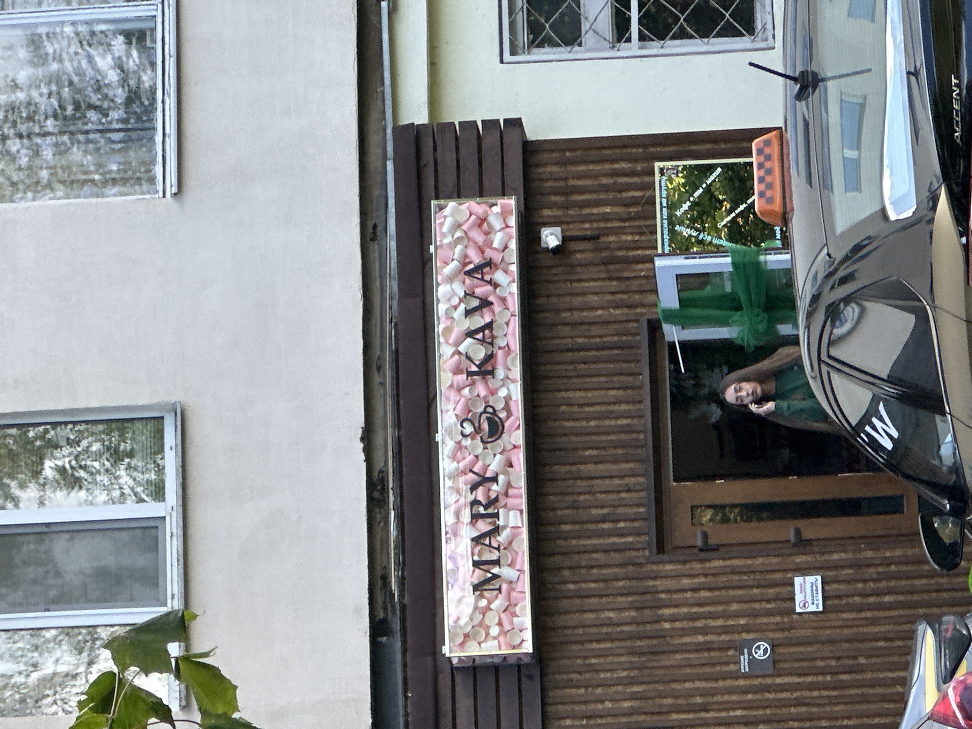 The image captures the facade of a cafe named "MARY KAVA" in Minsk, Belarus. The cafe's sign is decorated with an assortment of small pink and white disposable cups. A woman with long dark hair, wearing a green top, stands in the doorway, engaged in a phone conversation. In the foreground, the roof of a black taxi is visible, featuring an orange and black checkered taxi sign. Behind the taxi, parts of two cars are visible. The overall setting appears to be a street in an urban environment, with buildings and windows visible in the background.