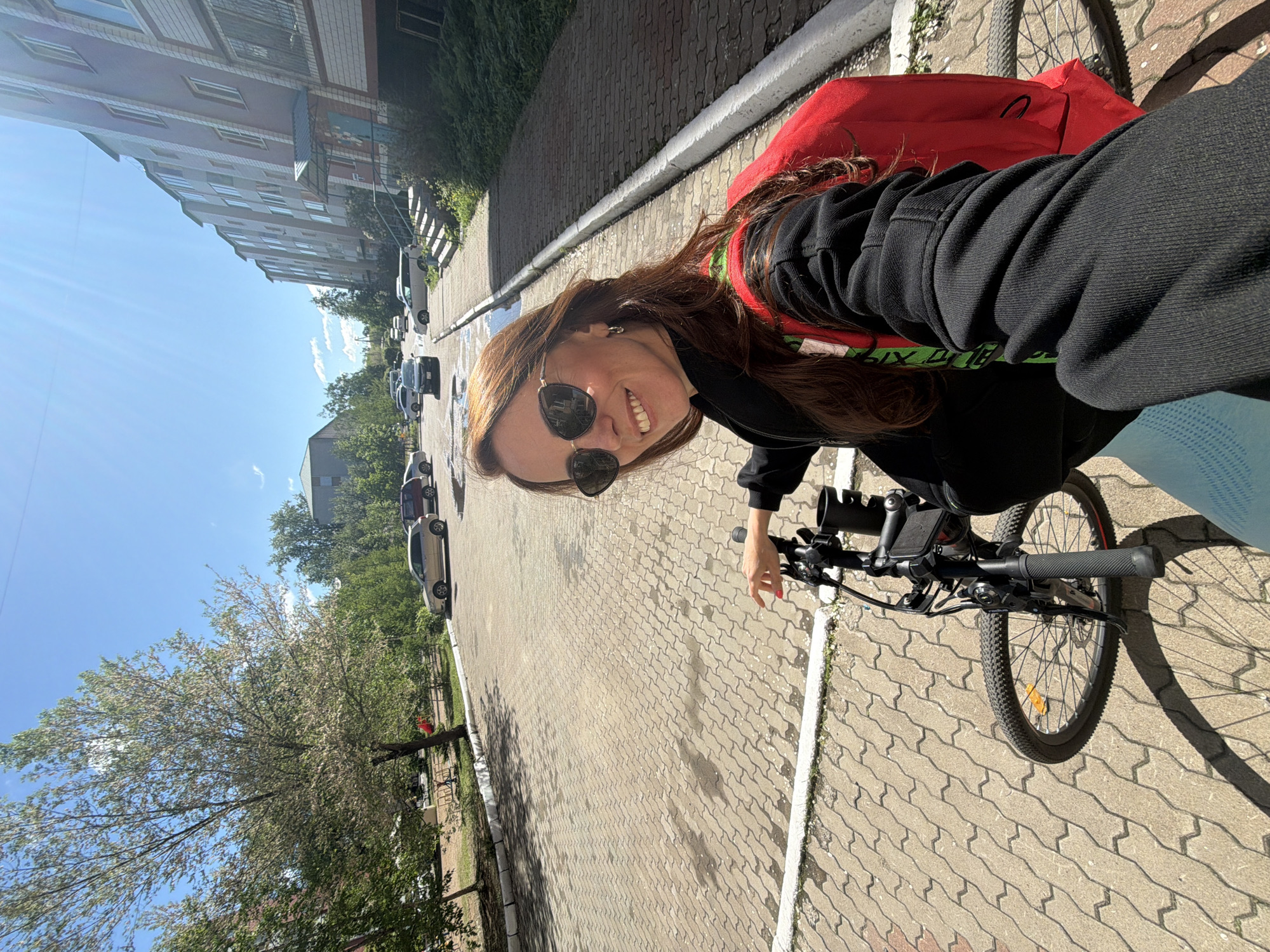 A woman is captured in a selfie, smiling broadly while on a bicycle on a sunny day in Birobidzhan, Russia. She has long, dark brown hair, wears sunglasses, and is dressed in a black hooded sweatshirt. A bright red backpack is slung behind her, with a glimpse of a light blue item, possibly clothing, visible beneath it. Her right hand rests on the handlebar of a black mountain-style bicycle.
The setting is an outdoor paved area, likely a sidewalk or path, constructed from interlocking concrete pavers. In the background, a long, multi-story residential building with a reddish and grey brick-patterned facade and numerous windows stretches along the street. Cars are parked neatly along the curb. Beyond the vehicles, green trees, some appearing to bloom with white flowers, line the area. The sky is a clear, vibrant blue with a few scattered white clouds.
The strong shadows cast on the pavement suggest it is daytime, possibly mid-morning or early afternoon. The woman's cheerful expression indicates a pleasant and active mood. Partially visible green Cyrillic text, possibly "АТЛХ", can be seen on the red backpack, though it is largely obscured and difficult to read.