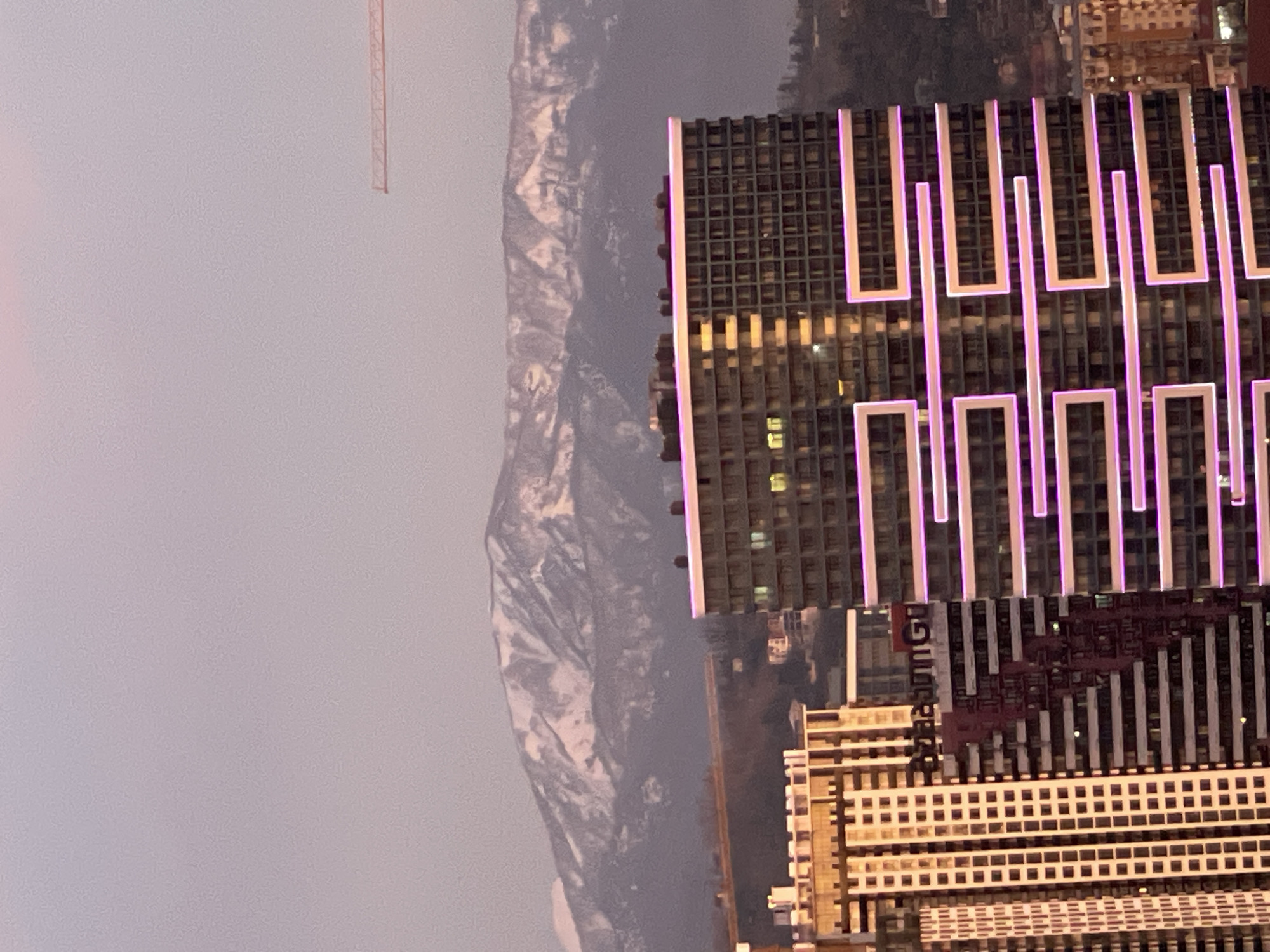 The image shows a nighttime view of Batumi, Georgia, featuring a snow-dusted mountain range partially obscured by fog or haze in the background. In the foreground, there are several high-rise buildings. One prominent building is modern in design, featuring a grid-like exterior with illuminated pink or purple vertical bars that create a decorative pattern. Adjacent to this building is another, taller building with a dark facade, with a decorative pattern on the middle section. A crane is visible in the upper-left corner. The sky is overcast.