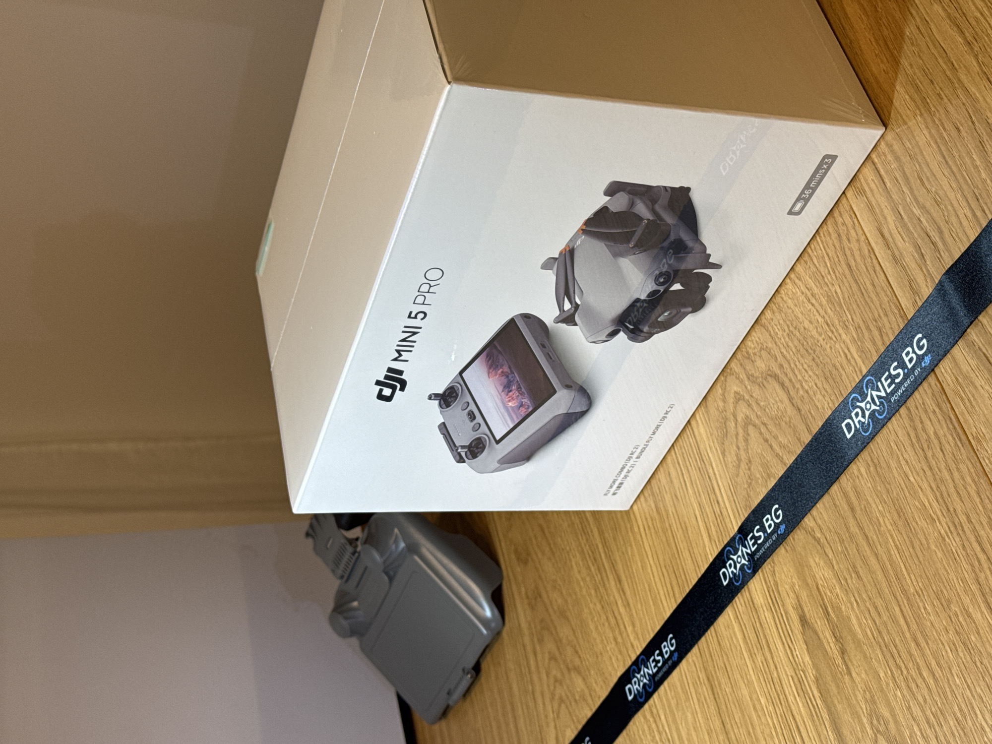 The image shows a white box for a DJI Mini 5 Pro drone, partially visible on a light brown wooden surface. Next to the box is a black lanyard with "DRONES BG POWERED BY DJI" printed in white. A gray drone battery is visible on the left side of the image. The image was likely taken indoors. The location is Sofia, Bulgaria as the lanyard says DRONES.BG.