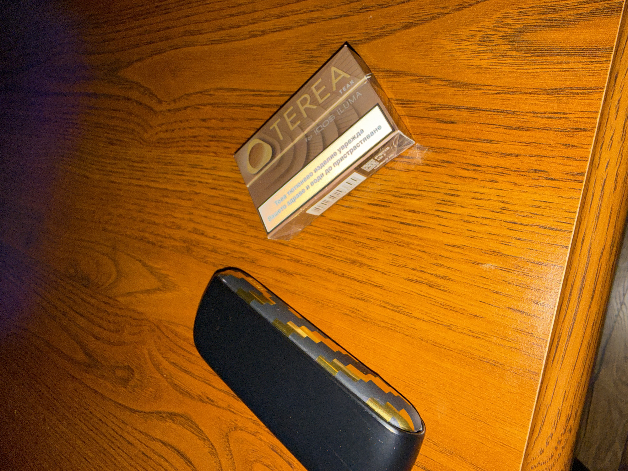 The image shows a pack of TEREA Teak for IQOS ILUMA and an IQOS device on a wooden surface. The TEREA pack is positioned diagonally in the upper center of the frame. Below it, towards the left, is the IQOS device, also diagonally placed. The wooden surface has a linear grain pattern, and the lighting appears to be artificial, potentially from an indoor setting. There are no people visible. The OCR text on the TEREA pack is in Bulgarian, which suggests the image may have been taken in Bulgaria, possibly in a city like Sofia.