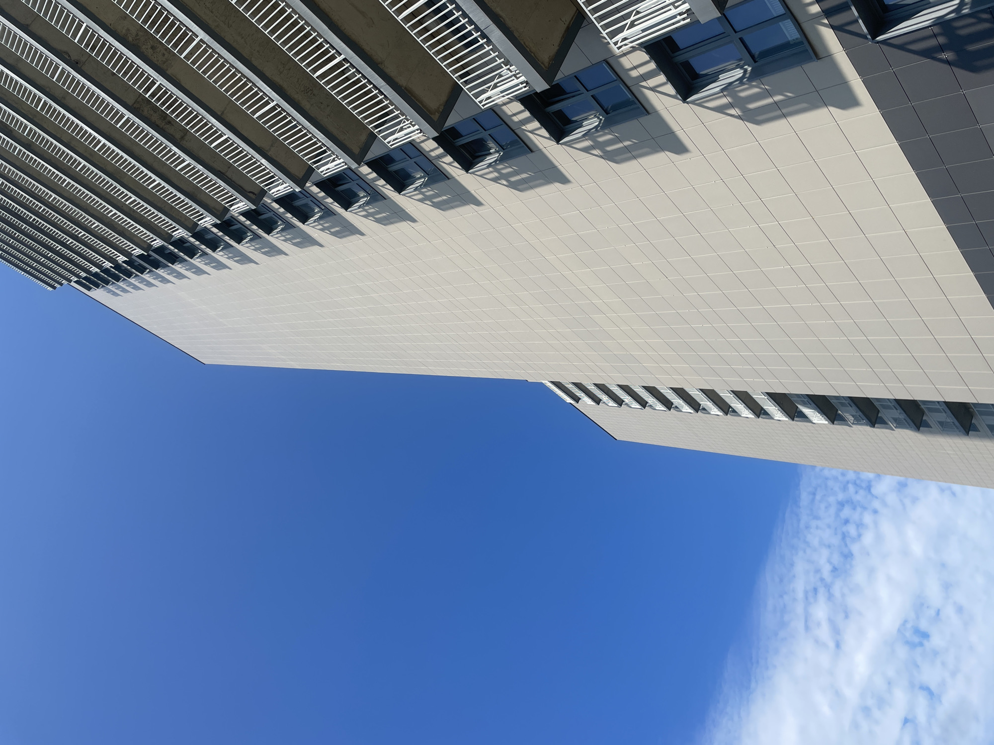 An upward-angled exterior shot of a modern, light-colored high-rise apartment building in Ladozhskoe Ozero, Russia, against a clear blue sky with a few wispy white clouds in the lower left corner. The building features repetitive balconies with white railings and dark-framed windows that cast distinct shadows on the building's facade. The facade is composed of square tiles, with a darker band of tiles near the bottom. No people are visible in the image.