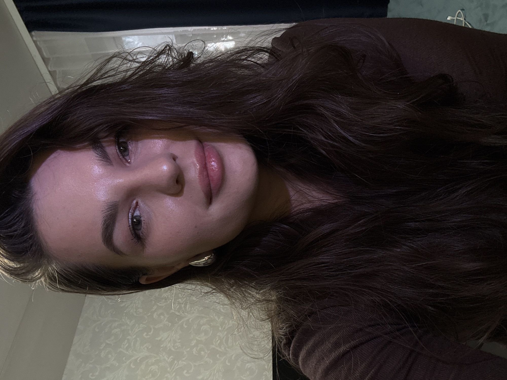 A young woman with long, dark, wavy hair is pictured from the chest up, looking directly at the camera. She is wearing a dark brown, ribbed top. She has a light-colored earring in her left ear. Her face is illuminated by a pinkish light, casting a shadow on the left side of her face. The background appears to be an indoor setting with a patterned wallpaper on the left and dark curtains and a window on the right. There is no indication of the city of Chaikovsky, Russia, in the image.