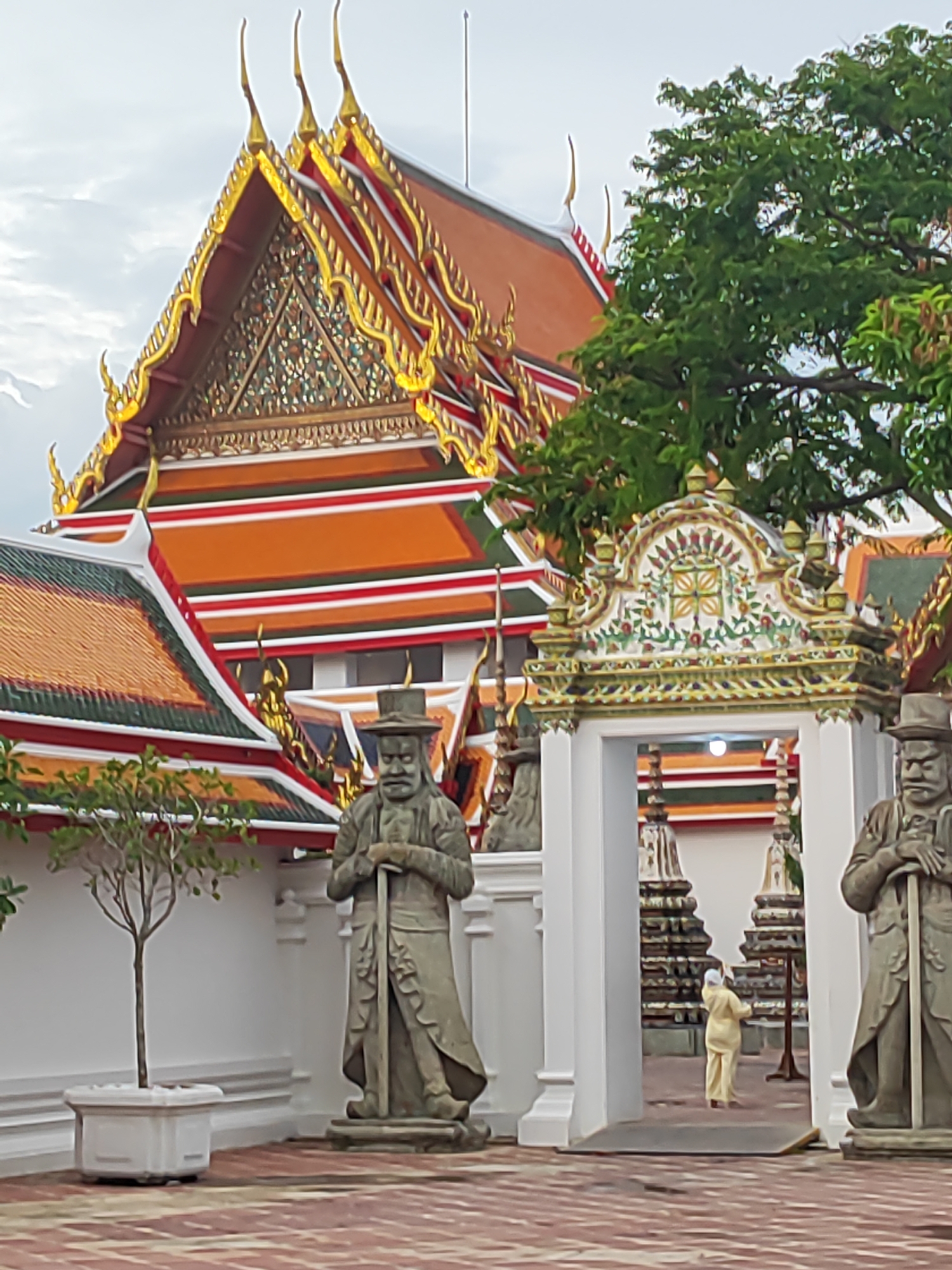 The image depicts the grounds of Wat Pho, a Buddhist temple in Bangkok, Thailand. In the foreground, two large, weathered stone statues of guardians stand on either side of a decorative archway. The guardians are depicted wearing hats and holding staffs. Beyond the archway, a person dressed in light yellow traditional clothing walks away from the camera, heading towards the temple structures. The temple buildings feature ornate, multi-tiered roofs with vibrant orange tiles and gilded detailing, characteristic of Thai temple architecture. Lush green trees partially obscure the upper portions of the temple. The ground is paved with reddish-brown bricks. The sky above is overcast, with hints of light blue. A small, potted tree stands to the left of the left guardian.