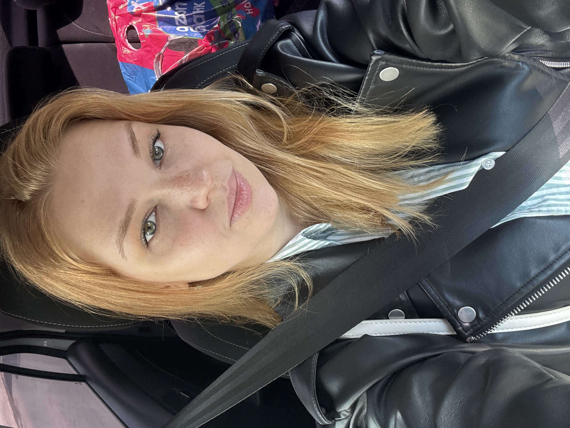 A young woman with blonde hair and green eyes is seen from the chest up, looking at the camera. She is wearing a black leather jacket over a striped shirt and a seatbelt is visible across her chest. She is seated in a car, as indicated by the car interior visible in the background. To her right, a colorful shopping bag is partially visible. The scene is set in the city of Zavolzhye, Russia.