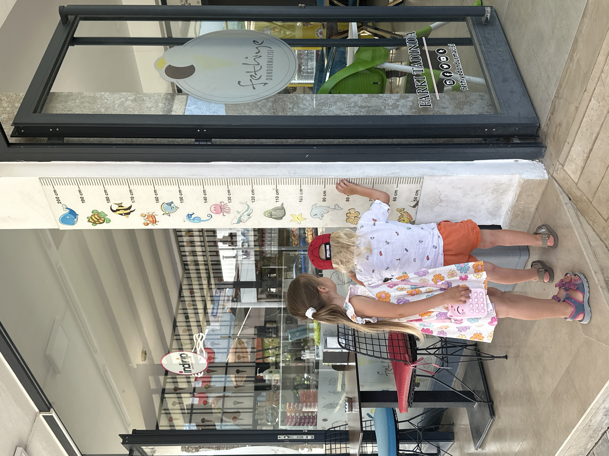 Two young children, a girl and a boy, are standing and measuring their heights against a height chart attached to a wall outside a shop in Fethiye, Turkey. The girl has long brown hair in a ponytail, wearing a floral dress and sandals. She's holding a small pink purse. The boy, wearing a red "SPIDER-MAN" hat and an orange short is standing next to her. The shop's glass front displays signage, including the shop's name, "Fethiye Dondurmacisi," along with the phrase "Farkı Tadinda" and social media icons. The chart has cartoon sea animals.