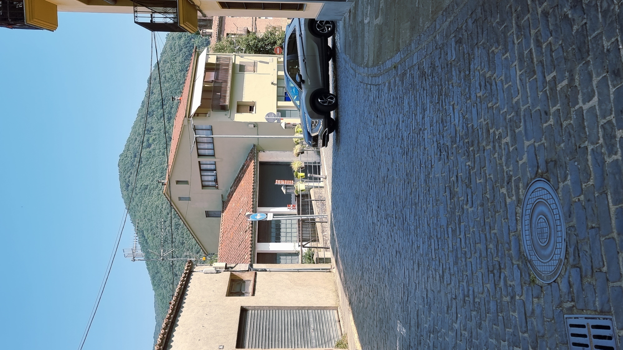 The image depicts a street scene in Sant Joan Les Fonts, Spain, characterized by a cobblestone road and surrounding buildings. A modern, dark grey SUV is positioned on the right side of the road, partially obscuring the view of a narrow, winding street. The vehicle appears to be stationary or moving slowly.
In the background, several buildings with terracotta tile roofs are visible. One building on the left features a large, metallic garage door. Another building in the center has multiple windows and a balcony with an awning. A smaller structure with a tiled roof and rolling metal doors is situated between these two.
The street itself is paved with dark grey cobblestones, with a circular manhole cover and a grated drain visible in the foreground. A blue and white circular traffic sign is posted on a pole near the center of the image.
The environment suggests a clear, sunny day, with a vibrant green, tree-covered mountain in the distant background. Power lines crisscross the sky. The overall impression is that of a quiet, European town or village.