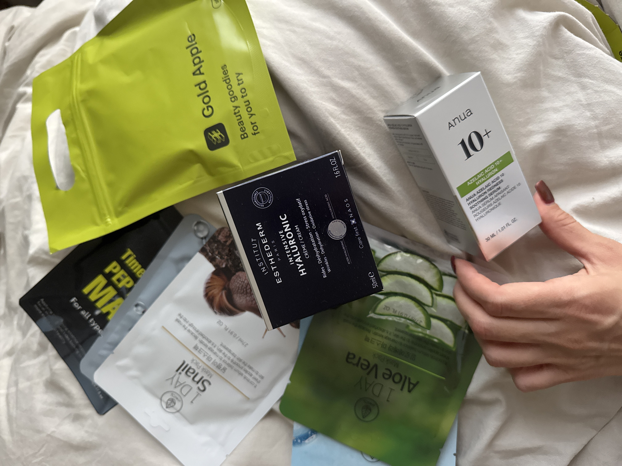 The image shows an assortment of skincare products arranged on a white bedsheet. A person's hand is holding a white box of "Anua 10+ Azelaic Acid 10 Hyaluron Soothing Serum." The other products include a green "Gold Apple" bag, a black "Time Pep MA" mask, a white "1 Day Snail Mask Pack," a black box of "Institut Esthederm Paris Intensive Hyaluronic Crème," and a green "1 Day Aloe Vera Mask Pack." The environment is clean and domestic. There is no evidence to suggest the location of the image is in Dubai, United Arab Emirates.