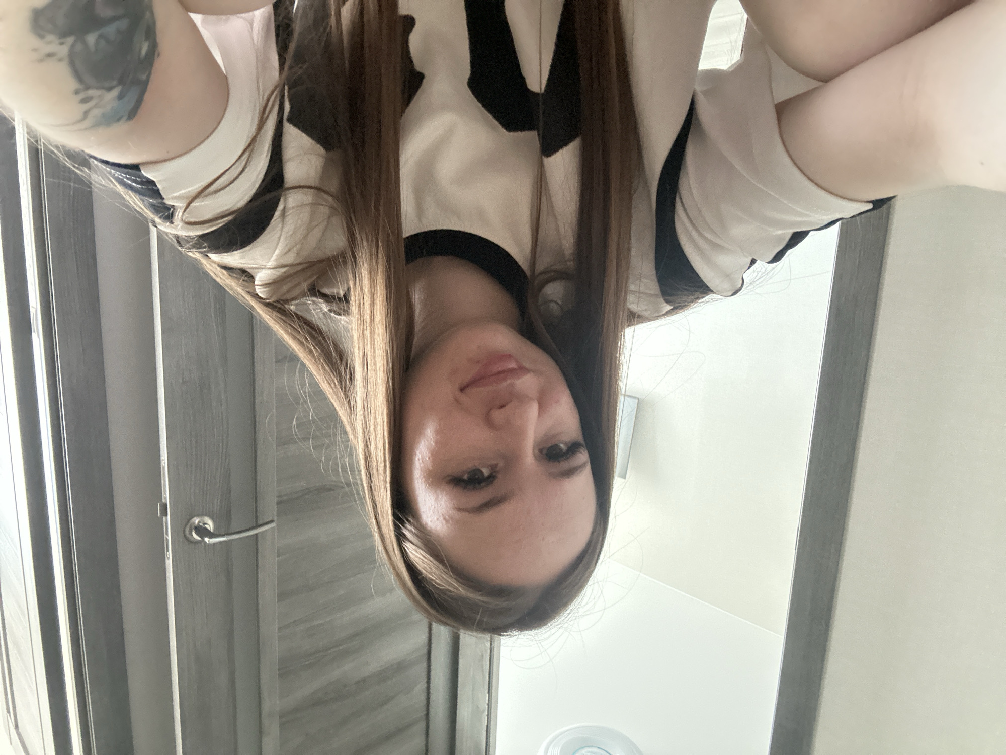 An upside-down selfie features a young woman looking directly into the camera. Her long, straight, light-brown hair hangs downwards, framing her face. She has fair skin, with some minor blemishes visible on her forehead and chin. Her dark eyes are open, and her lips are slightly parted, forming a neutral or subtle expression.
She is wearing a light-colored (possibly white or off-white) top with bold black patterns or accents on the shoulders and collar area. A tattoo, appearing to be dark blue and black and possibly depicting a floral or abstract design, is visible on her upper left arm.
The background suggests an indoor setting. On the left side of the frame, a closed door with a silver handle is visible. The door appears to be made of a dark gray or wood-textured material. The right side of the frame shows a plain, light-colored wall. A circular object, possibly a light fixture or vent, is partially visible towards the bottom right corner of the image. No text is visible in the frame.