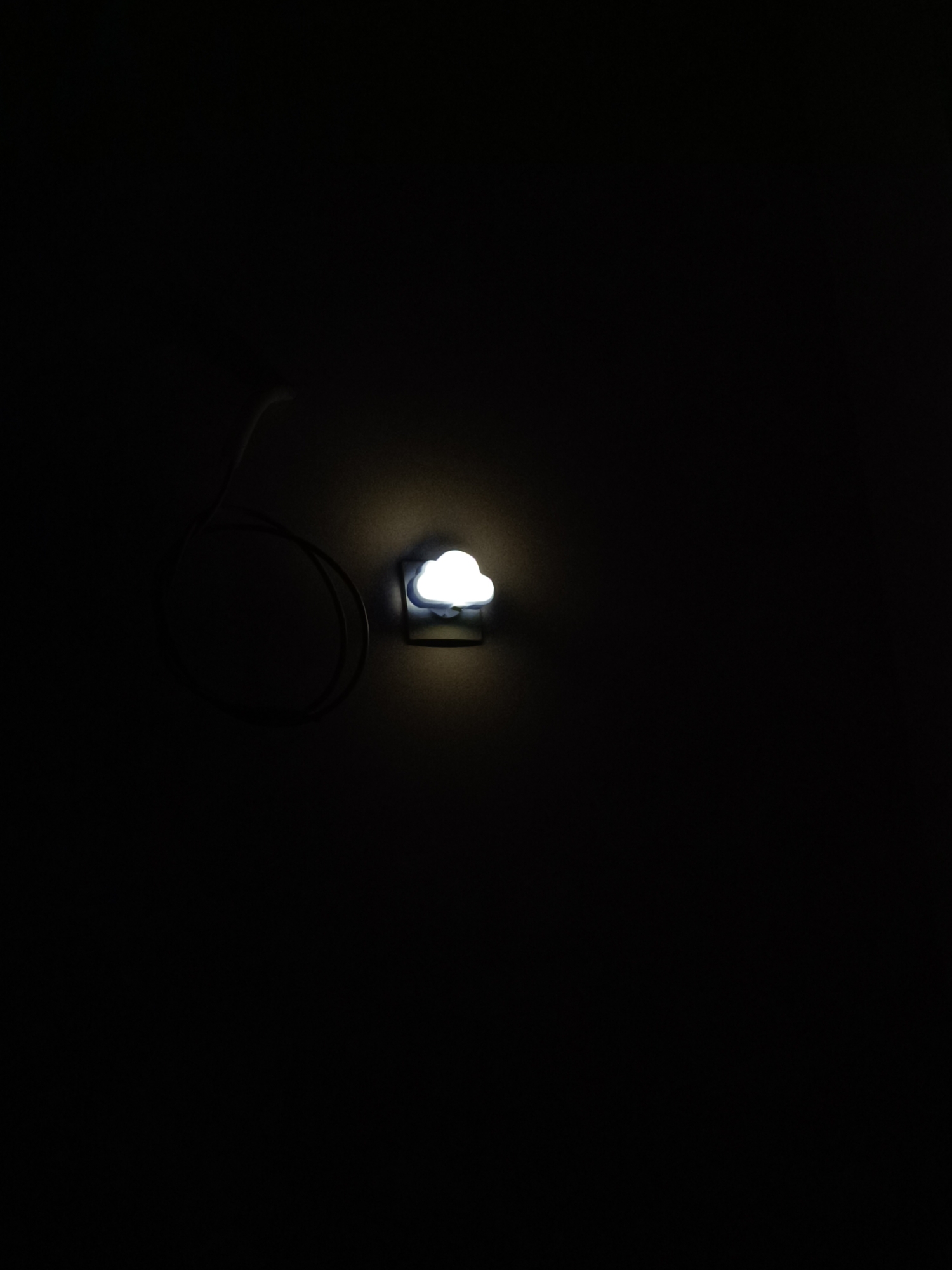 A small, cloud-shaped nightlight affixed to a dark, presumably interior wall is illuminated. A power cord extends from the nightlight, curling loosely on the wall. The surrounding environment is predominantly black, suggesting darkness and a lack of ambient light.