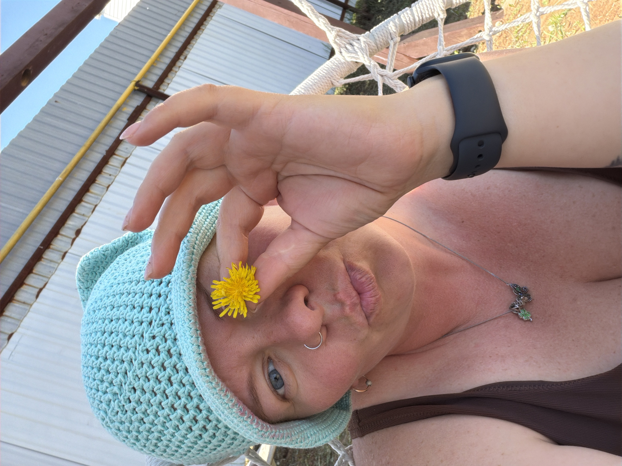 A woman wearing a blue crocheted hat is holding a yellow dandelion flower up to her eye, partially obscuring her face. She has a silver nose ring, a silver necklace with a four-leaf clover charm, and a dark grey smartwatch. She is wearing a brown top. The background includes part of a hammock, a corrugated metal structure with a yellow metal bar, and some green foliage. The photograph appears to be taken outdoors, in daylight. The location is Chaikovsky, Russia.