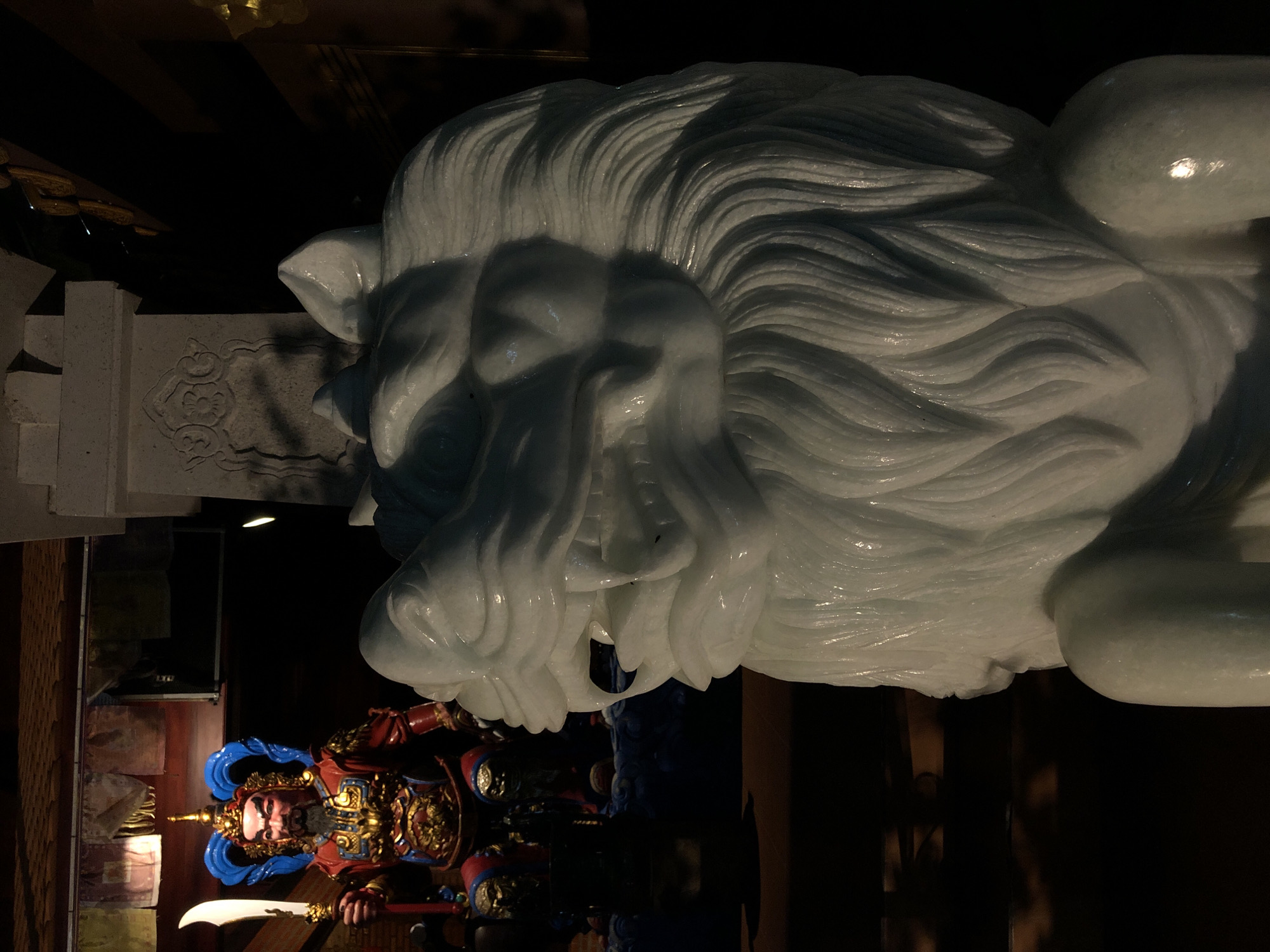 This image shows a close-up of a white, carved stone lion sculpture, likely a foo dog, with a dark background. The sculpture is detailed, with intricate carvings representing the lion's mane and facial features. In the lower left portion of the image, a statue of a warrior figure in ornate armor is visible. This warrior statue is depicted with a red, blue, and gold costume, a black beard, and is holding a curved sword. The lighting suggests an indoor setting, possibly a temple or cultural display, with the shadows indicating a dimly lit environment. The overall mood is somber and majestic. There is no visible text in the image. The setting is identified as Mũi Né, Vietnam.