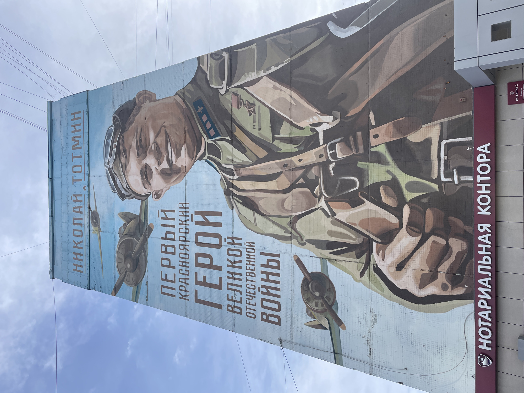 The image captures a large, vertically oriented mural painted on the side of a building, depicting a male military figure, likely a pilot or soldier. The man is shown from the chest up, wearing a flight helmet with goggles pushed up, and a military uniform adorned with medals and insignia on his chest and shoulder. He has a determined, slightly smiling expression. In the background of the mural, two propeller-driven fighter aircraft are visible, suggesting an aviation theme.
The mural features prominent Russian text. On the left, it reads "НИКОЛАЙ ТОТМИН" (Nikolai Totmin). Below the figure's face, the text states "ПЕРВЫЙ КРАСНОЯРСКИЙ ГЕРОЙ ВЕЛИКОЙ ОТЕЧЕСТВЕННОЙ ВОЙНЫ" (First Krasnoyarsk Hero of the Great Patriotic War), indicating the mural commemorates a local hero from Krasnoyarsk, Russia, who fought in World War II.
Adjacent to the mural, on the right side of the image, is a modern building section with a dark red sign displaying "НОТАРИАЛЬНАЯ КОНТОРА" (Notary Office) in white Cyrillic letters. A smaller sign above it also reads "НОТАРИУС" (Notary). The scene is set outdoors under an overcast sky, with multiple power lines or cables visible stretching across the top. The building's surface underneath the mural appears to be made of small, light-colored tiles.