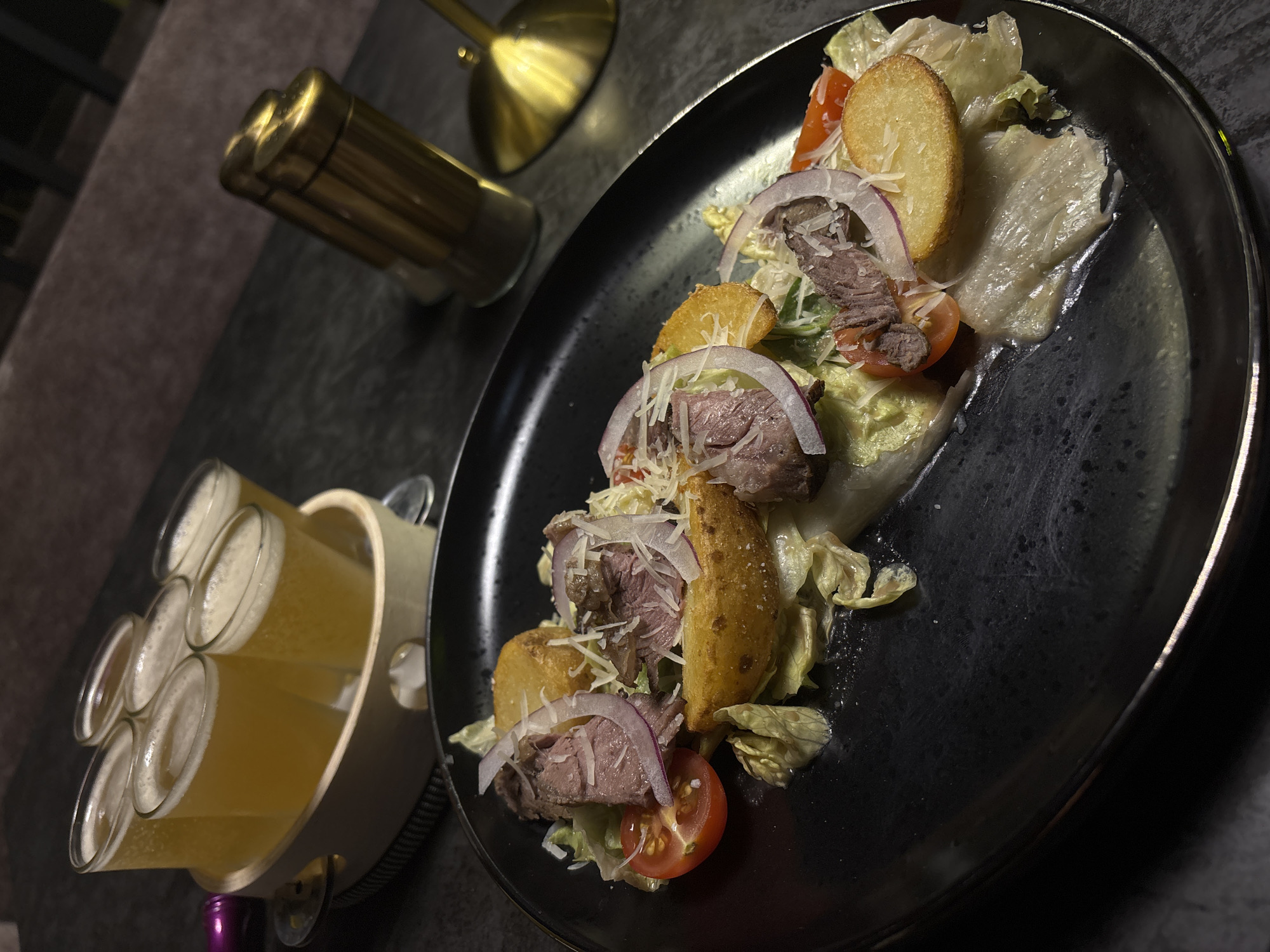 A dark, upscale establishment with dim lighting is the setting for this image. In the foreground, a black plate holds a meticulously arranged salad. The salad features sliced beef, lettuce, thinly sliced red onion, halved cherry tomatoes, and several golden-brown potato wedges. Shredded white cheese is sprinkled generously over the top. The plate itself is dark and slightly reflective.
To the left of the plate, a wooden carrier holds six small glasses, each filled with a frothy, pale yellow beverage, likely beer. The glasses are arranged in two rows of three.
In the background, a brass-colored shaker and another metallic object, possibly a cocktail shaker or a similar bar tool, are visible on a dark surface. The lighting casts reflections on these objects. The overall impression is one of a well-presented meal and drinks, suggesting a night out or a gathering in a bar or restaurant. No people are visible in the image, and there is no discernible text. The scene appears to be indoors, likely in the evening or at night due to the low light conditions.
