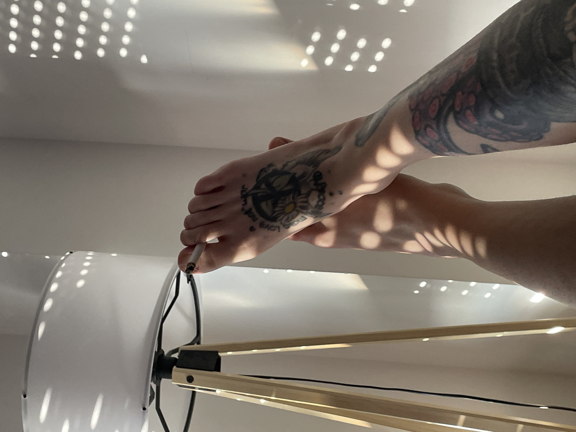 The image shows a close-up of a person's tattooed foot holding a lit cigarette near a modern-looking floor lamp with a large, white, circular shade. The person has tattoos on their foot and arm. The background consists of a white wall with dappled sunlight patterns. There is no indication that this is taken in Belgrade, Serbia.