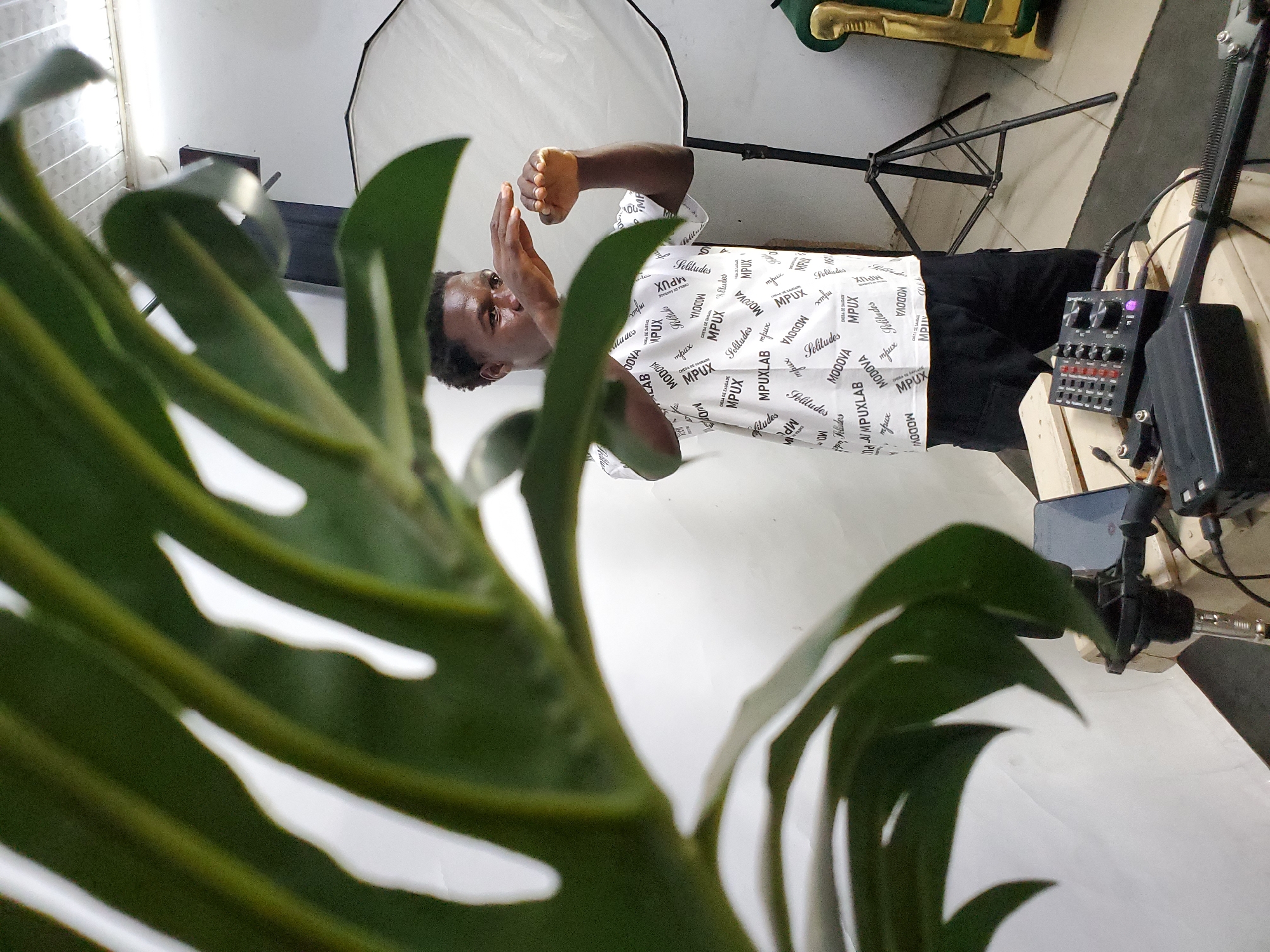 A medium shot depicts a man in a white patterned shirt and black pants, gesturing with his hands in a studio setting in Mararaba, Nigeria. A large, green monstera plant is in the foreground, partially obscuring the scene. Behind the man, there is a white backdrop illuminated by a softbox on a stand. An audio mixer and other recording equipment are visible on a table to the right. The orientation of the image is rotated clockwise by approximately 90 degrees.