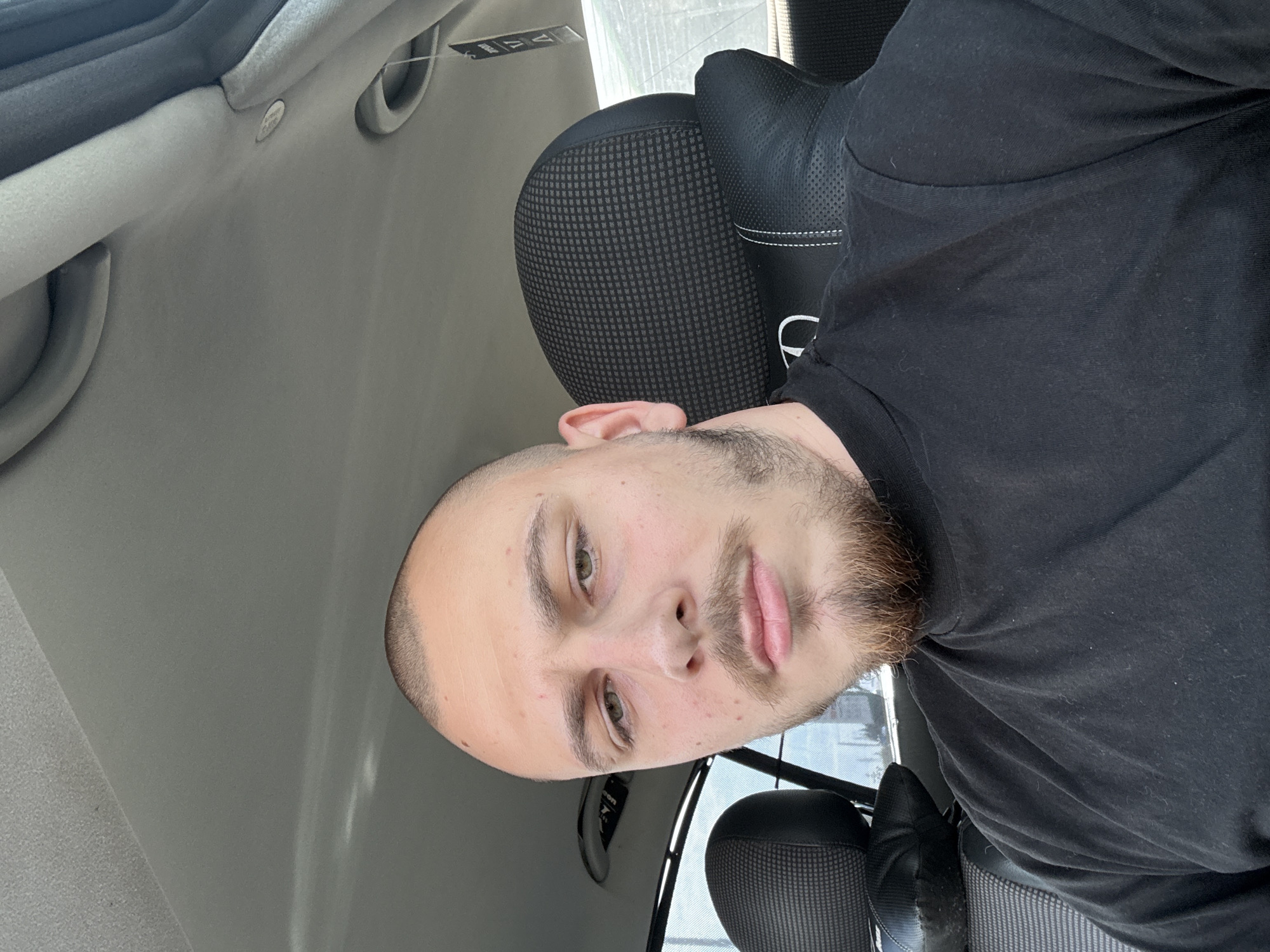 A man is in a car. He has fair skin, a shaved head, and a brown beard. He is wearing a black t-shirt. The car's interior is grey. The car's seat is black. The window shows a glimpse of the outside. Based on the context clues, the man is likely in Stara Zagora, Bulgaria.