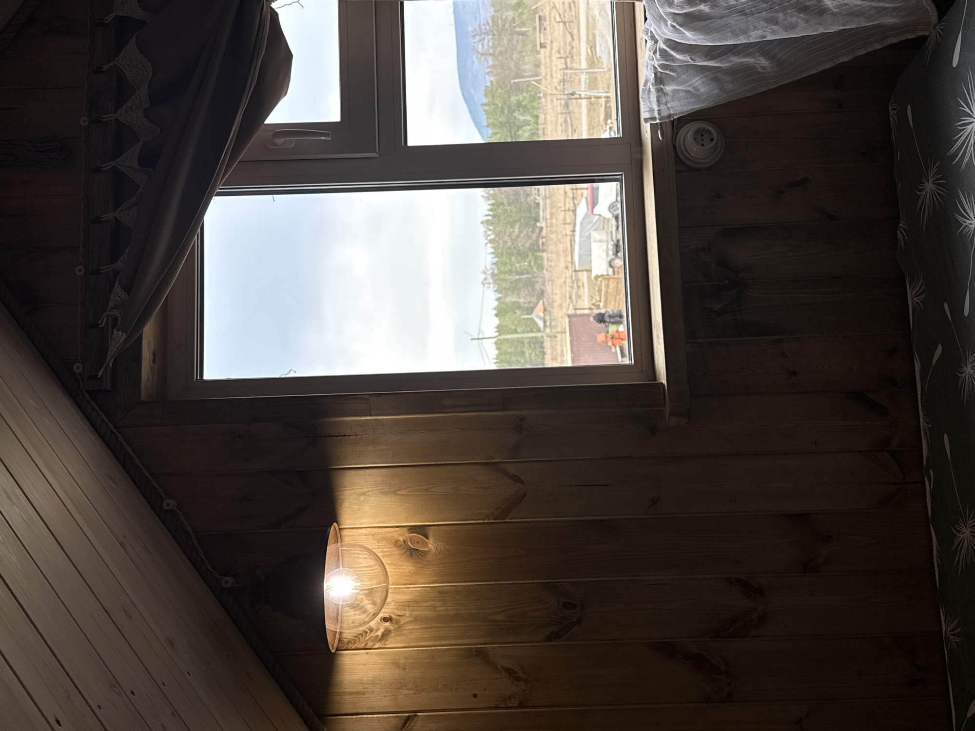 A photograph taken indoors depicts a window overlooking an outdoor scene. The window, situated in a wooden-paneled room with an angled ceiling, frames a view of Tyulyuk, Russia. Through the window, a landscape is visible, showing sparse vegetation and a distant, indistinct mountain range. A partially drawn curtain adorns the left side of the window, while a patterned curtain is present on the right. On the interior wall, a light fixture illuminates the wooden paneling. The overall lighting in the room is a mix of natural light from the window and artificial light from the fixture, creating a play of shadows on the wood.