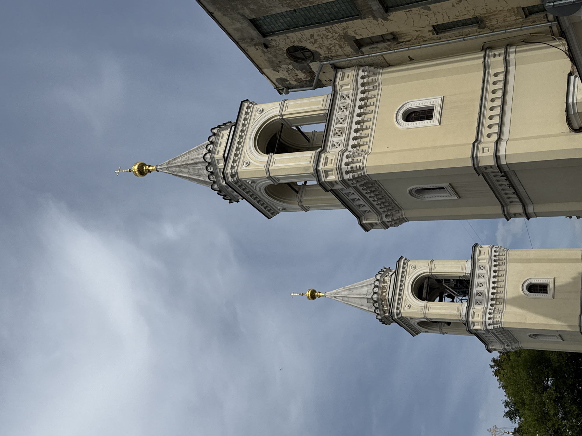 The image captures the bell towers of the Church of the Ascension of the Lord in the village of Kolomenskoye, Moscow, Russia. Two distinct bell towers are visible, the taller one in the foreground and a slightly shorter one to the left. Both are painted a pale yellow and feature tiered designs with arched openings, decorative cornices, and conical roofs topped with golden crosses. The foreground tower has bells visible within its arched openings. To the right of the bell towers, a weathered, older building with peeling paint and dark-paned windows is partially visible. The sky above is overcast with grey clouds, though patches of brighter sky are visible. Green trees are present behind and to the left of the bell towers. The perspective is looking upwards at an angle, emphasizing the height of the structures against the sky.
