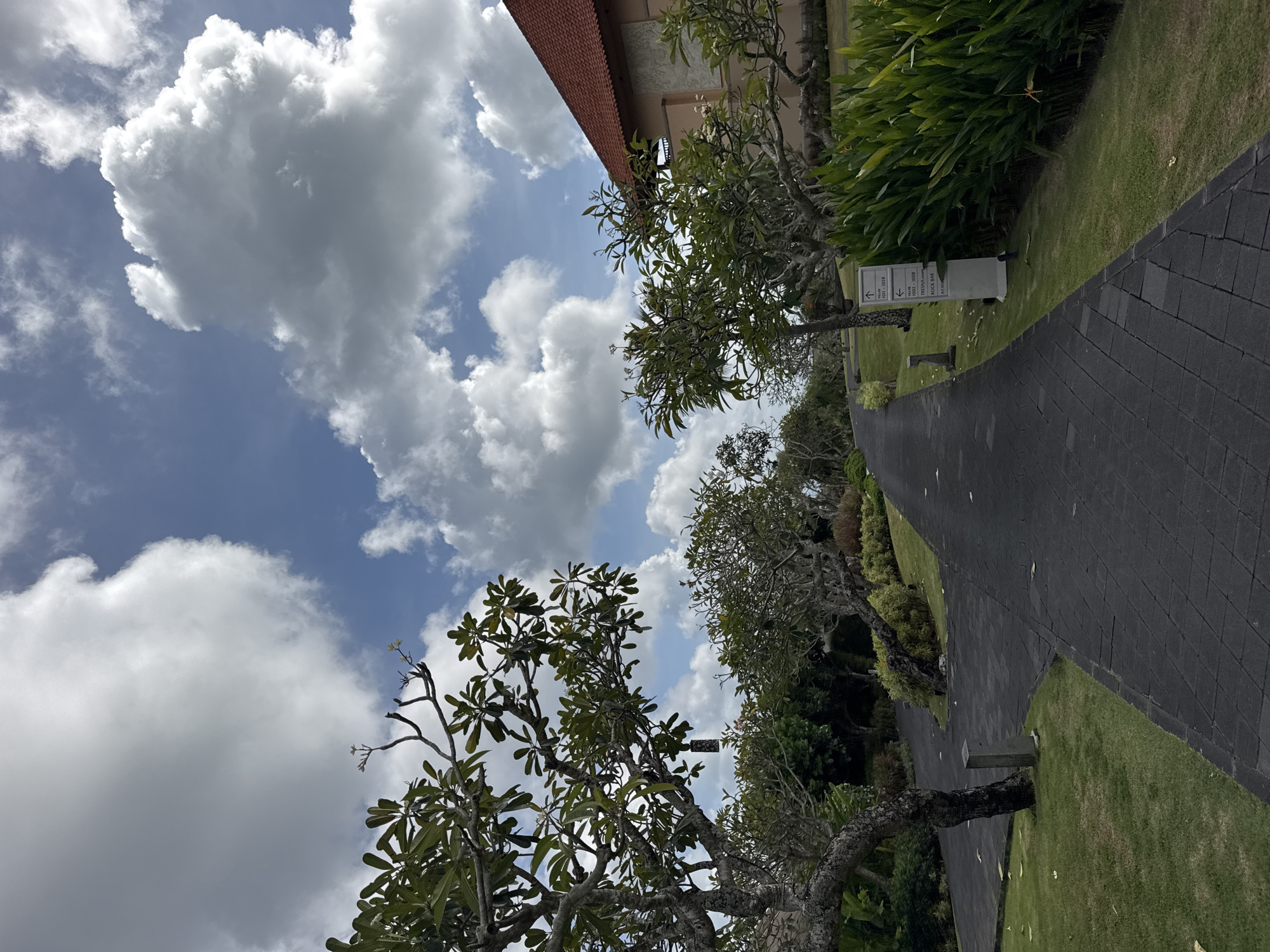 The image shows an outdoor scene in Kuta Selatan, Indonesia. A dark grey paved walkway extends into the distance, bordered by green grass and various trees and plants. The sky is visible above, featuring a mix of blue and white, with puffy clouds scattered throughout. Part of a building with a red roof is visible at the top of the frame. A sign is present near the walkway, possibly providing directions or information.