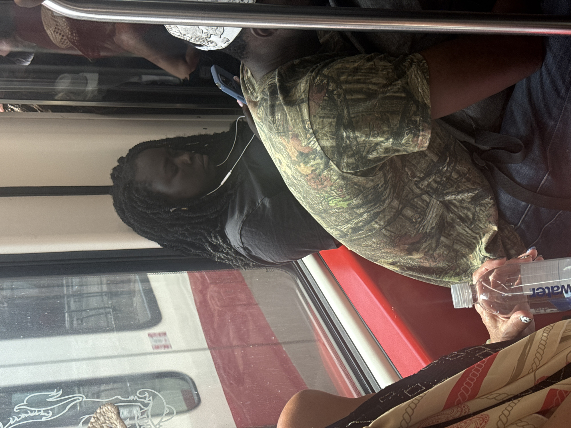 The image captures a candid scene inside a Calgary Transit CTrain car. A woman with dark skin and braided hair is seated, wearing earbuds and looking down at her phone. In front of her, another person wearing a camouflage-patterned shirt leans forward, also seemingly engaged with a phone. A plastic water bottle with the label "Water" is visible in the foreground, held by a person whose hand with decorative nail art is resting on it. The red seats and window of the CTrain are discernible in the background, with a reflection of the train car and the exterior visible through the glass. The lighting suggests it is daytime.