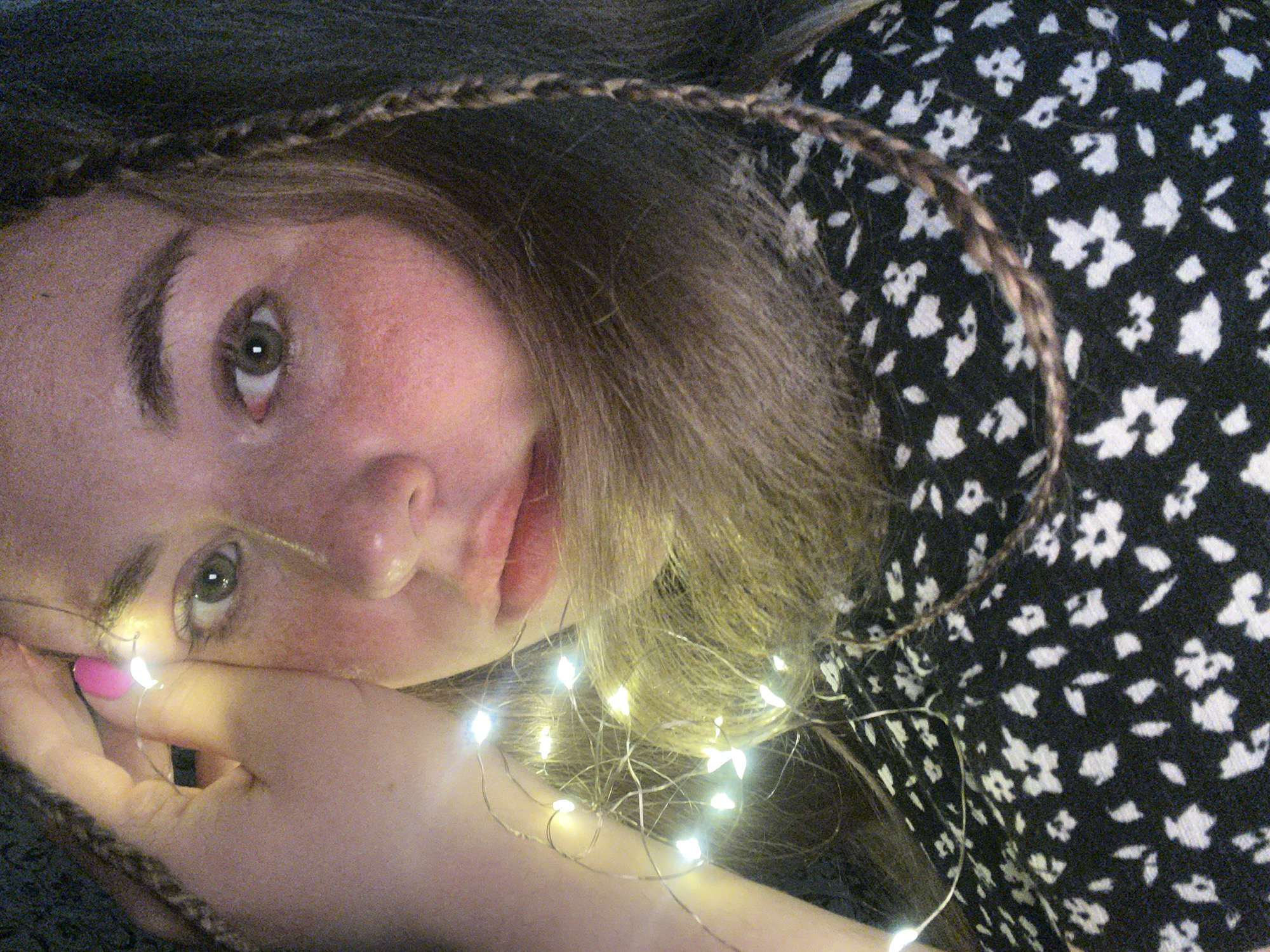 A young woman is close-up, posing with fairy lights. Her face is angled slightly upwards, with green eyes looking towards the camera. Her complexion is fair, and she has medium-length light brown hair with a braid on the top and side. She is wearing a black and white floral print dress. One of her hands is positioned near her face, with pink nail polish. Illuminated fairy lights are wrapped around her hand and hair. The background is dark and out of focus. There is no visual information to support that the image was taken in the city of Chaikovsky, Russia.