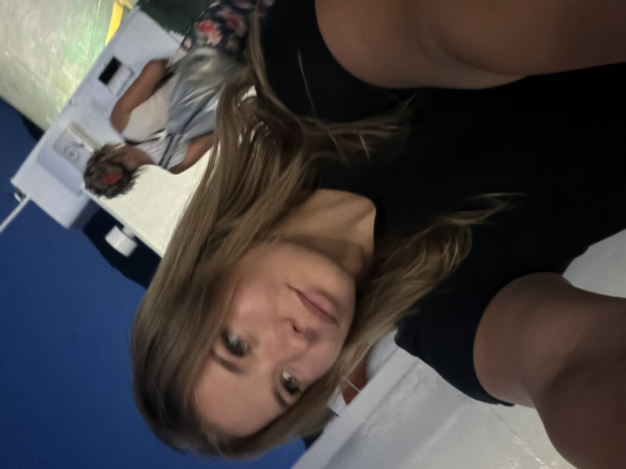 The image is a selfie taken at night, possibly on a ferry or boat, with a dark blue sky visible in the background. The main subject is a young woman with long blonde hair and light-colored eyes, looking towards the camera. She is wearing a black t-shirt. In the background, another person with short, light-colored hair and a white top is visible, along with a bag with a floral pattern. The setting appears to be outdoors, with some structures or railings visible. There is no indication of the location being in Đurići, Montenegro.
