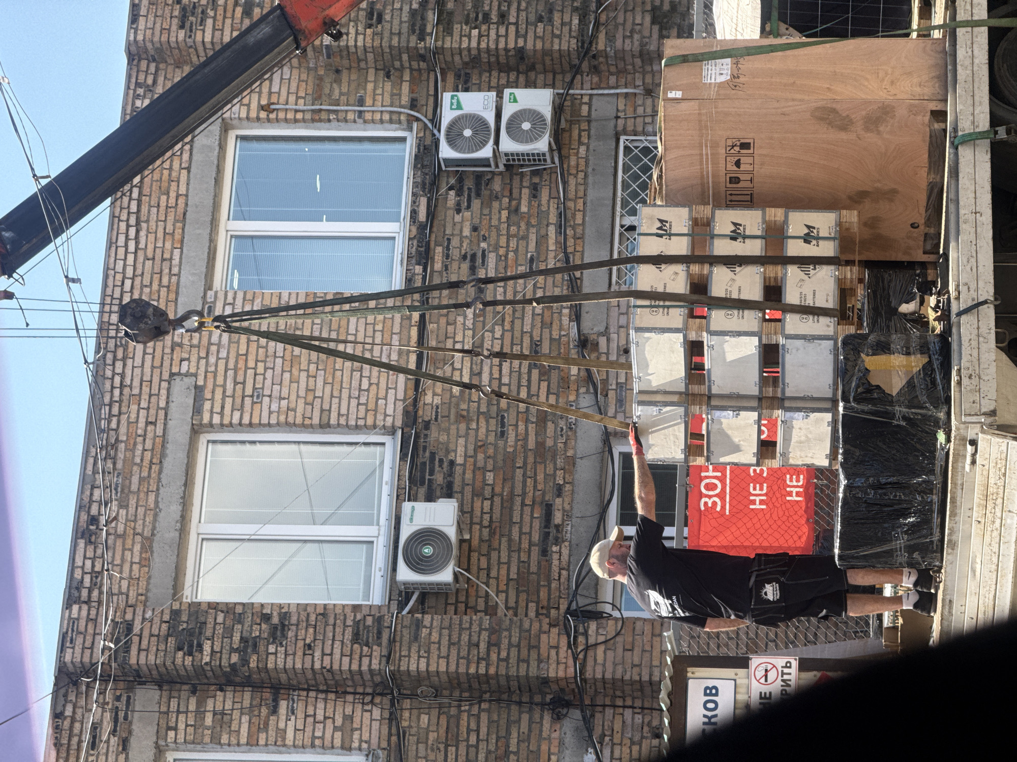 The image depicts an outdoor scene in front of a brick building. A crane is lifting a pallet of wooden crates, secured by straps, which are positioned on the back of a flatbed truck. A man wearing a black t-shirt, black shorts, and a light-colored baseball cap is standing on the truck, reaching towards the crates with his right hand. He is wearing red gloves. Several air conditioning units are mounted on the exterior of the building. Signs with Cyrillic lettering are visible on the left side of the image, one of which has a red background with white text. A large wooden crate is also on the truck to the right of the smaller crates. The sky above is clear and blue. The scene appears to be in an industrial or commercial area.