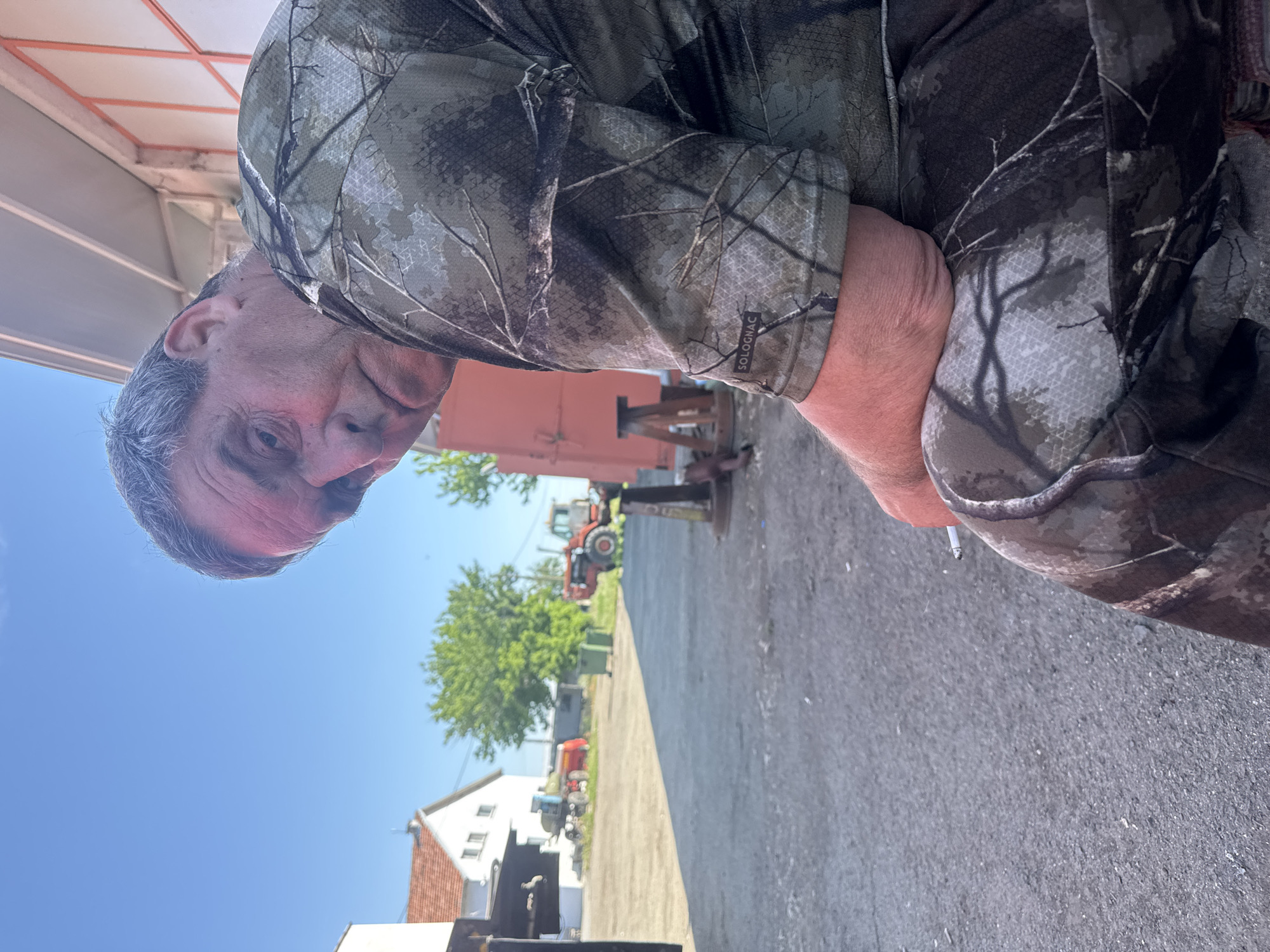 A middle-aged man is looking at the camera in what appears to be an outdoor scene. The man is wearing a camouflage shirt. The background shows a gray concrete ground, buildings, tractors and greenery under a clear blue sky. The man is holding a cigarette. The location is Sarnevo, Bulgaria.
