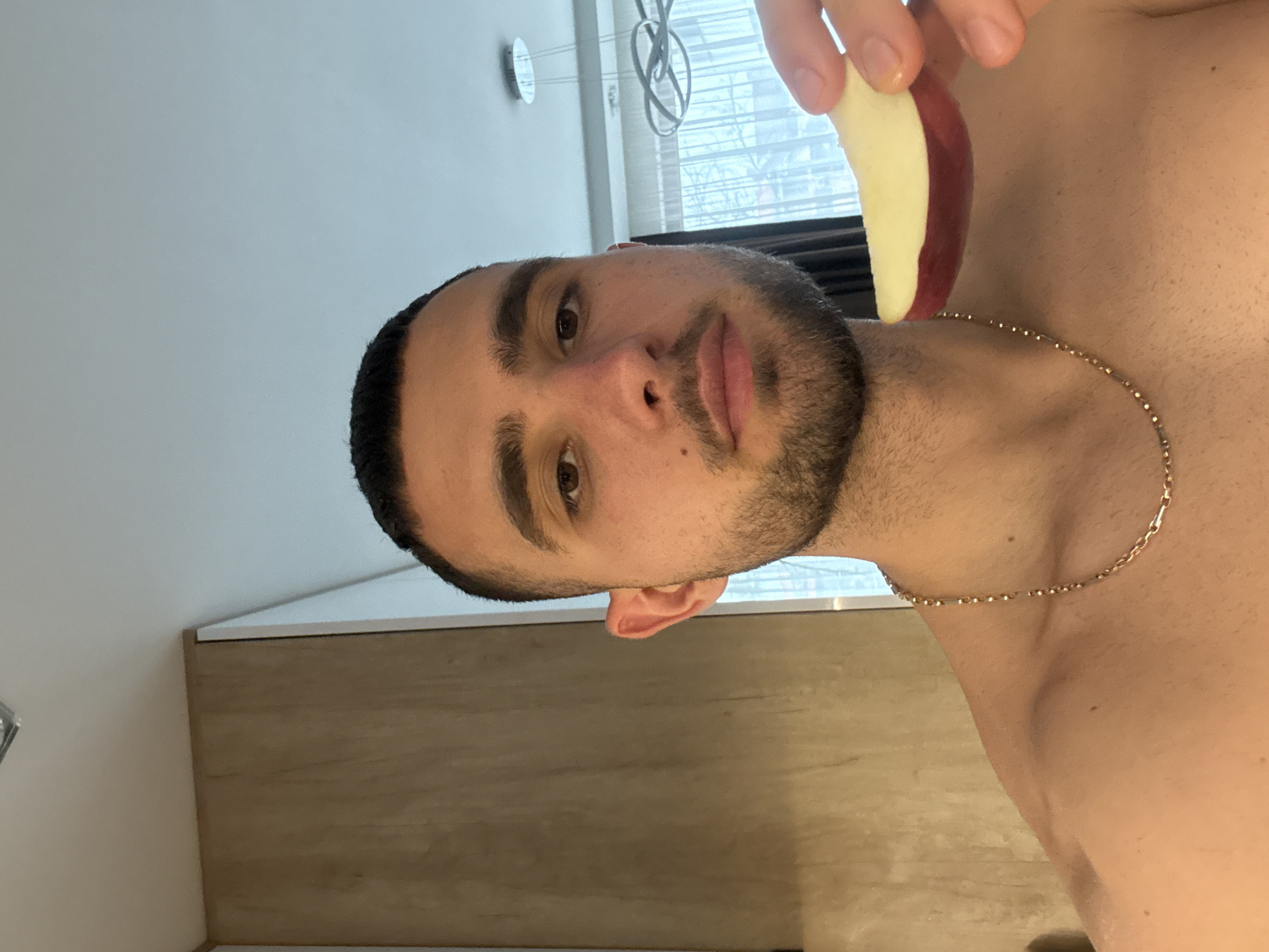A shirtless man with a beard is holding a slice of apple in his hand. He is wearing a gold chain. He is standing indoors near a window. Behind him, there is a cabinet and a light fixture. There is no indication of Gabrovo, Bulgaria.