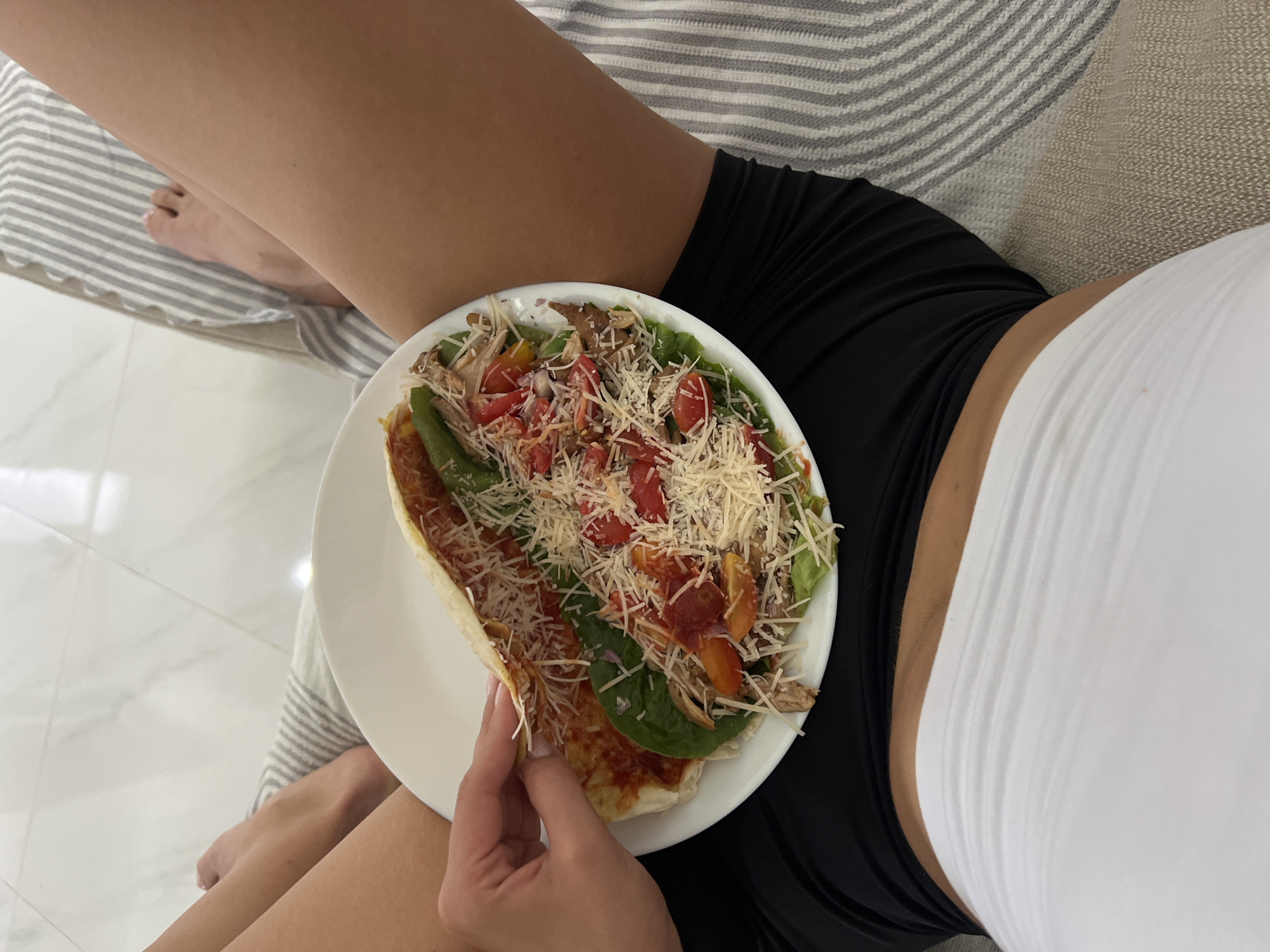A close-up shot shows a person holding a plate with an open-faced taco. The taco is filled with shredded meat, cherry tomatoes, lettuce, green peppers, and grated cheese. The person's hand, with light pink painted fingernails, is gently holding the edge of the taco. The person is wearing black shorts and a white bandeau top. They are seated on a striped couch. The background consists of a tiled floor and the couch. The scene is indoors.