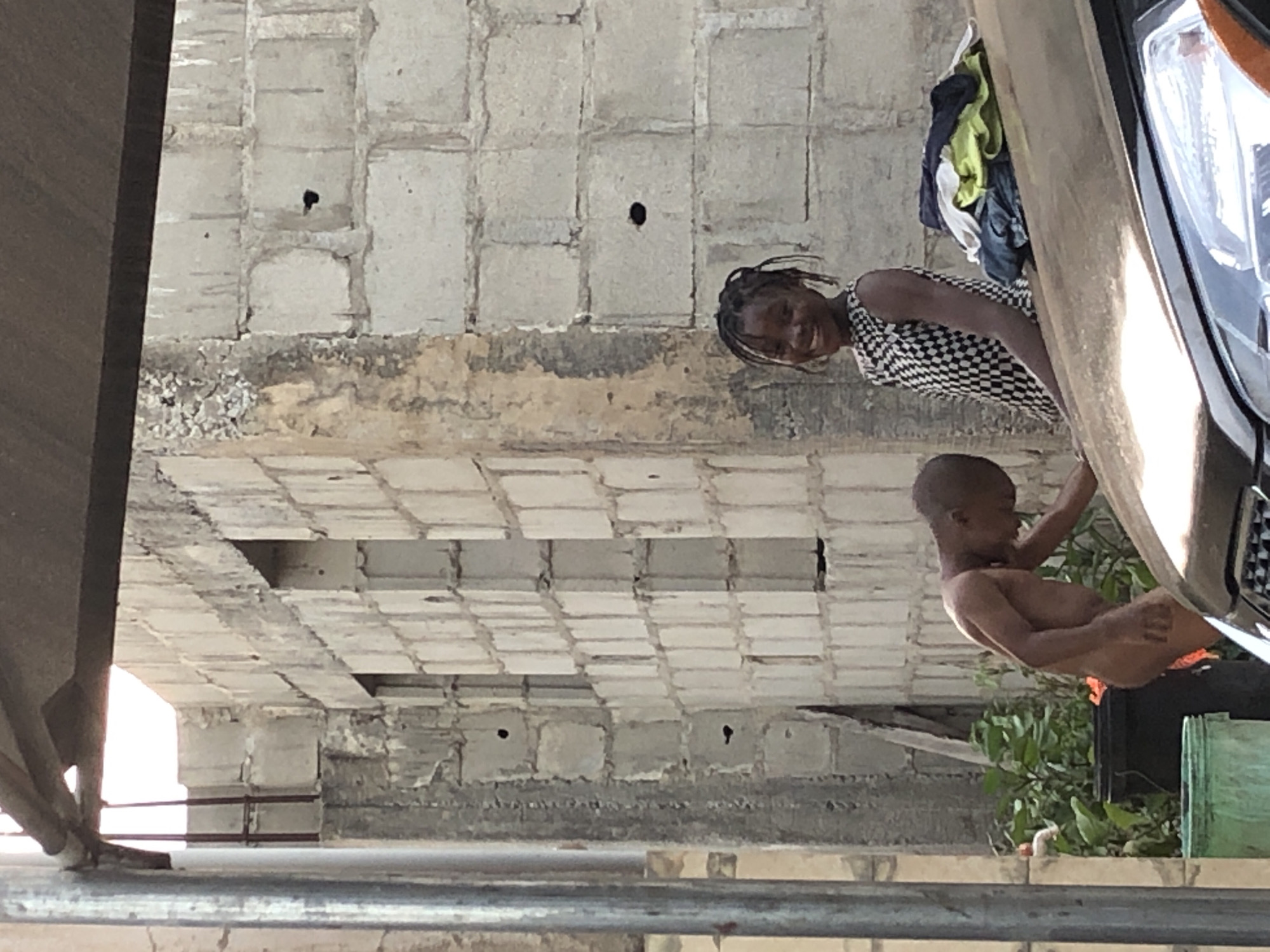 Captured from a low angle, the image portrays an urban scene. A woman with braided hair, clad in a black and white checkered top, smiles at the viewer. Next to her, a shirtless child is positioned near the front of a vehicle. Behind them, the backdrop consists of a concrete structure with a tiled ceiling and a wall featuring a grid-like pattern punctuated by dark holes. Adjacent to the car, some green plants are visible. The environment suggests an urban setting, potentially underneath an overpass or bridge.