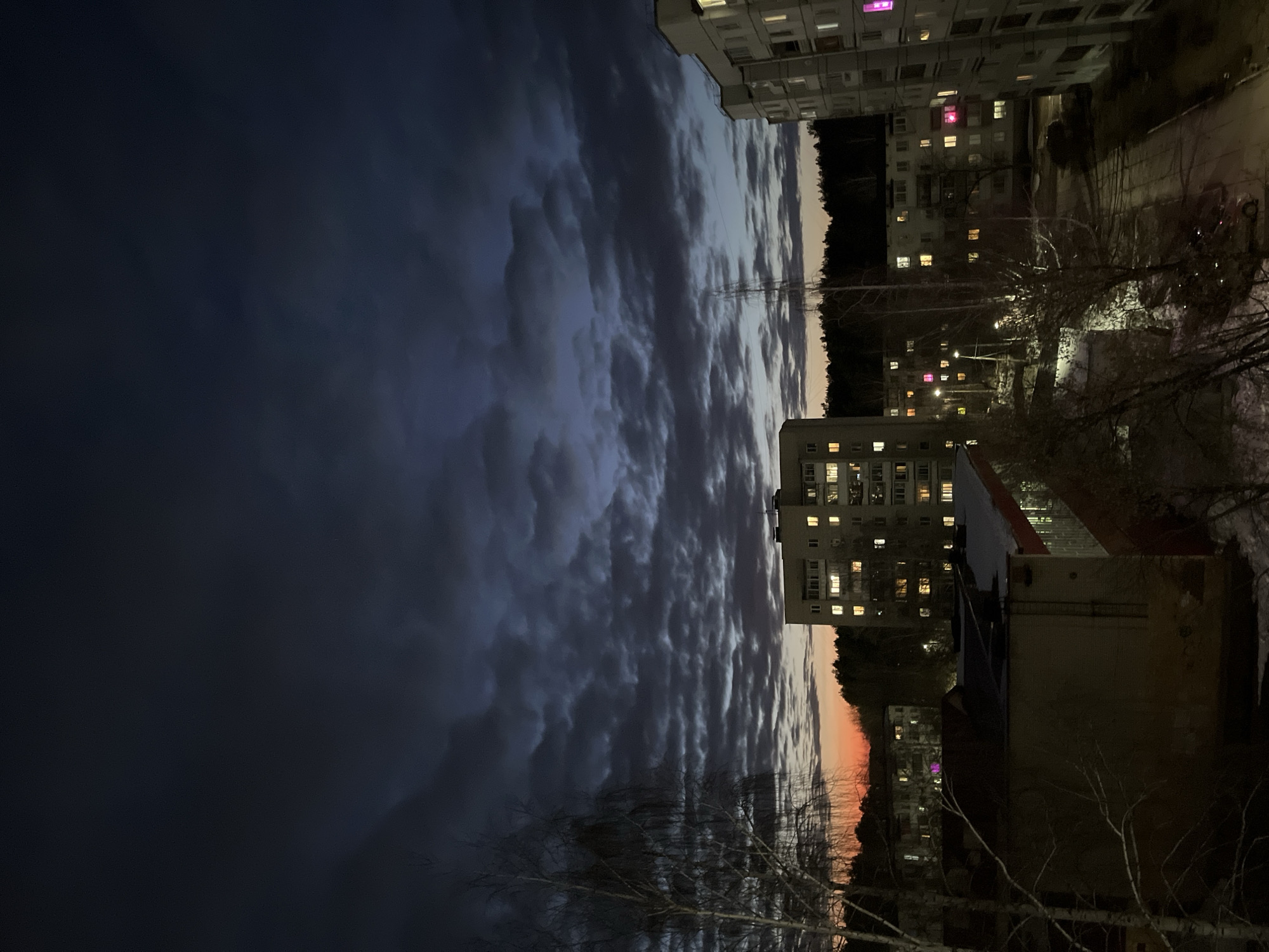 The image depicts an urban landscape in Chaikovsky, Russia, at dusk or early night. The sky occupies the upper two-thirds of the frame, characterized by dense, dark gray cloud formations with an underlying deep blue tone. A narrow band of orange-red light from the setting sun is visible along the lower horizon, transitioning upwards into the darker sky.
Multiple multi-story residential buildings are prominent across the midground and background. A large, light-colored apartment block stands near the center-left, featuring numerous windows, many of which are illuminated with warm yellow and white light. Another tall, light-colored apartment building is partially visible on the far right edge, also showing various lit windows, one displaying a distinct magenta glow. Between these larger structures, a line of dark coniferous trees forms a dense horizon, suggesting a forested area.
In the foreground and midground, numerous bare deciduous trees, possibly birches, exhibit fine branch structures without foliage. Patches of snow are visible on the ground and rooftops of some lower buildings in the foreground. A paved road or pathway, appearing dark and potentially wet or icy, is visible in the lower right quadrant, with a dark-colored sedan parked on its left side. Several streetlights are active, casting yellow illumination onto the scene. No individuals are visible within the frame.