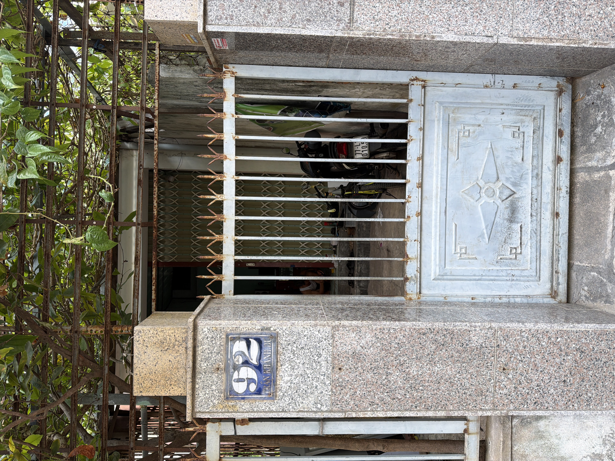 The image depicts the entrance to a residential property in South Quy Nhon Ward, Vietnam. A rusty metal gate, partially open, guards the entrance. The gate has a decorative pattern on its lower half and vertical bars above. To the left of the gate, a granite-clad pillar displays the house number "92" and the text "TRAN KHANH DU" on a blue tile. A motorcycle is parked just inside the gate. Overhanging the entrance is a metal structure covered with lush green foliage, suggesting a vine or tree providing shade. The overall scene conveys a sense of everyday life in a Vietnamese urban setting.