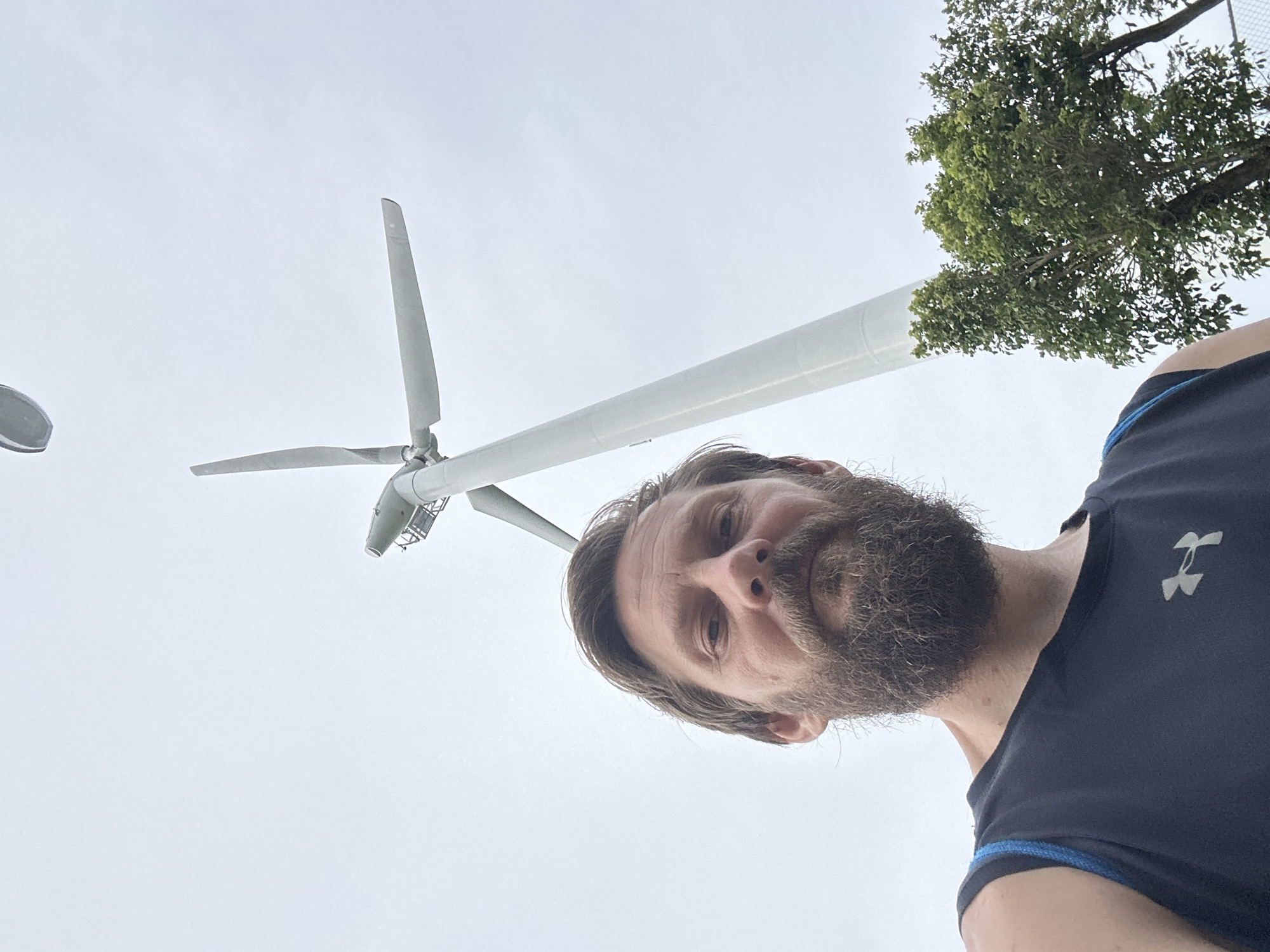 In Rawai, Thailand, a man with a beard and short hair is taking a selfie in front of a wind turbine. He is wearing a black sleeveless shirt with a blue strap over his shoulder. The wind turbine is tall and white, with three blades. There are also some green trees in the background, and the sky is overcast. The man's face is the primary focus of the image, with the wind turbine and surrounding environment providing context.