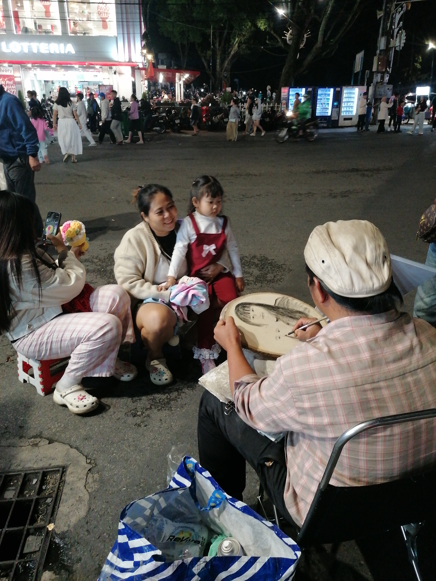 In the Vietnamese city of Da Lat, at night, an artist sketches on a circular wooden slab. A girl in a red dress sits with a woman as another woman captures a photo of a stuffed toy. The background features a brightly lit Lotteria restaurant and other street activity.