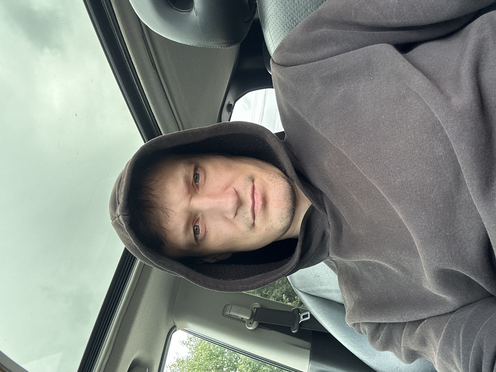 A man is in the driver's seat of a car. He is wearing a dark hooded sweatshirt. The car has a sunroof, which is open, showing a cloudy sky. The man is looking at the camera. The interior of the car appears to be dark leather. The background shows trees and greenery outside the car window. The location is not specified in the image or metadata.