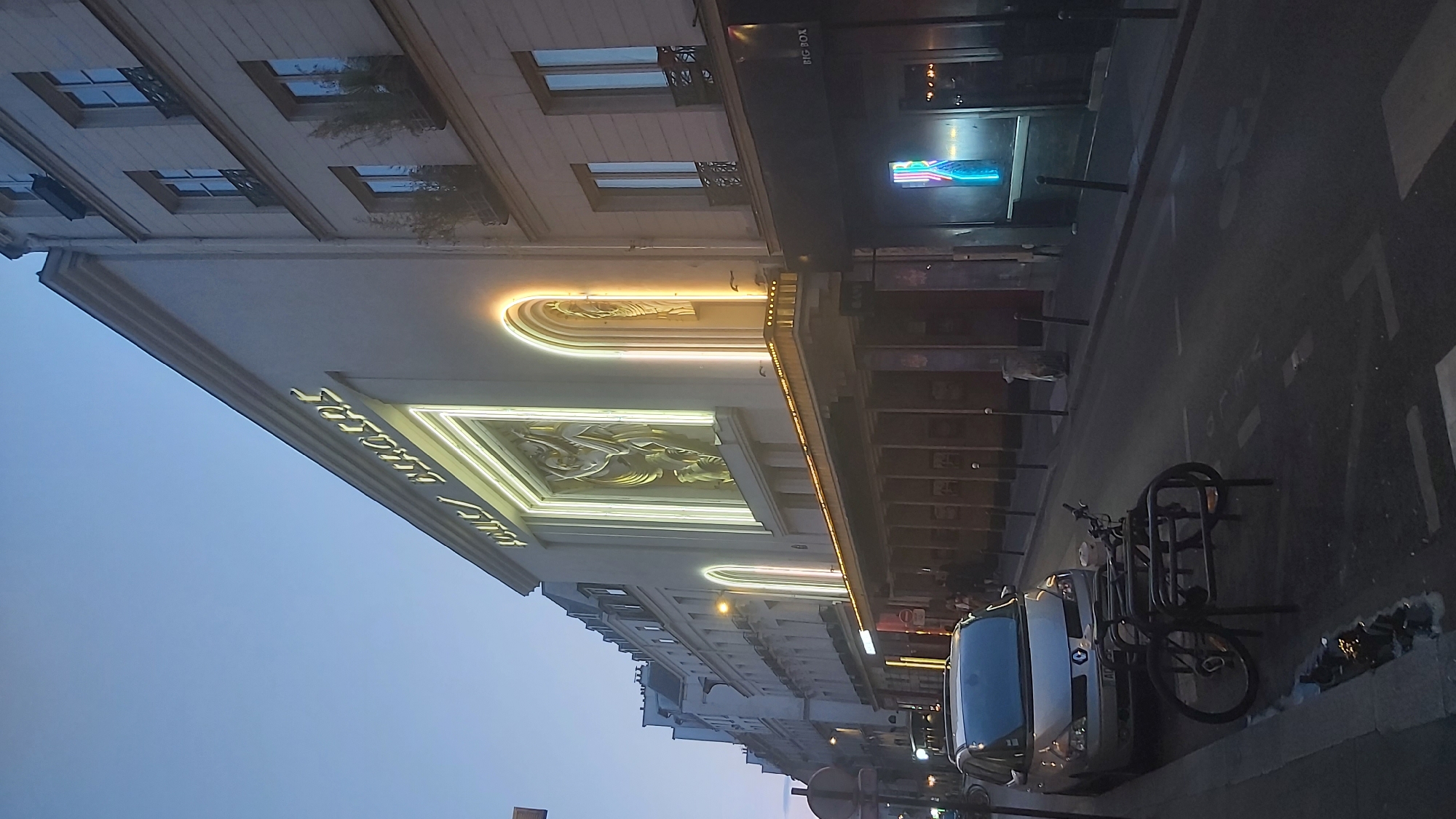 The image shows a view of the Théâtre Marigny on Avenue des Champs-Élysées in Paris, France, at dusk. The theater's facade is illuminated with soft, warm lighting around the marquee and the central artistic relief. To the left of the theater, a silver Renault Espace minivan is parked, and a bicycle rack with a bicycle is positioned in front of it. Further down the avenue, several other buildings with illuminated windows and signs are visible, suggesting an urban environment that is transitioning from day to night. The sky is a deep blue, characteristic of twilight. The street is paved and appears to be a typical Parisian boulevard, with streetlights and bollards lining the sidewalk. There are no people clearly discernible in the foreground, but some figures can be seen further down the street, indicating the presence of pedestrians. The overall atmosphere is calm and the scene is characteristic of a European city in the early evening.