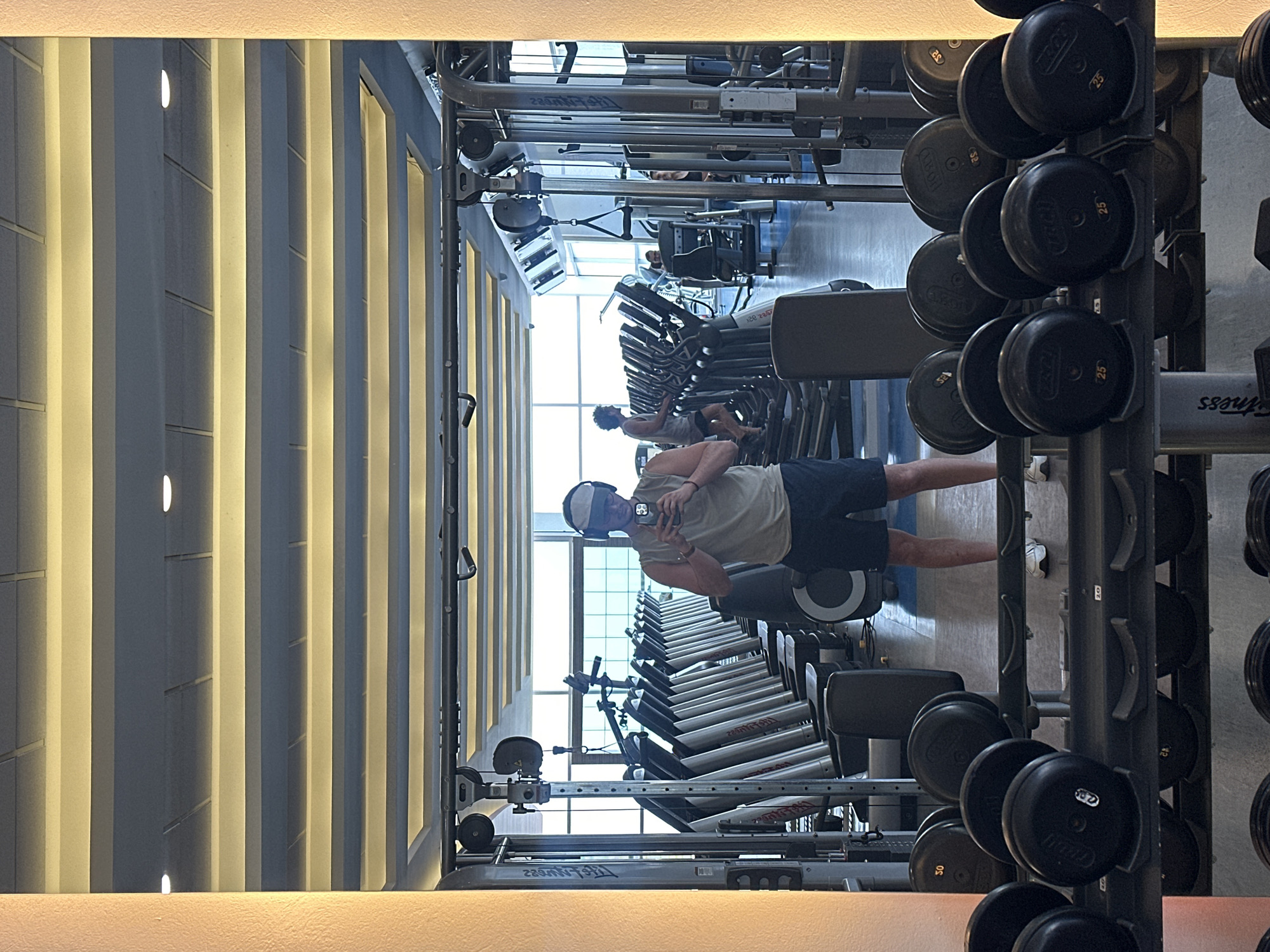 A person stands in a brightly lit gymnasium in Mexico City, Mexico, taking a selfie with their phone. The gym has rows of treadmills and weight racks. Other individuals are using the equipment. The lighting is a combination of natural light from windows and artificial lighting from overhead fixtures. The walls appear to be white or light gray.
